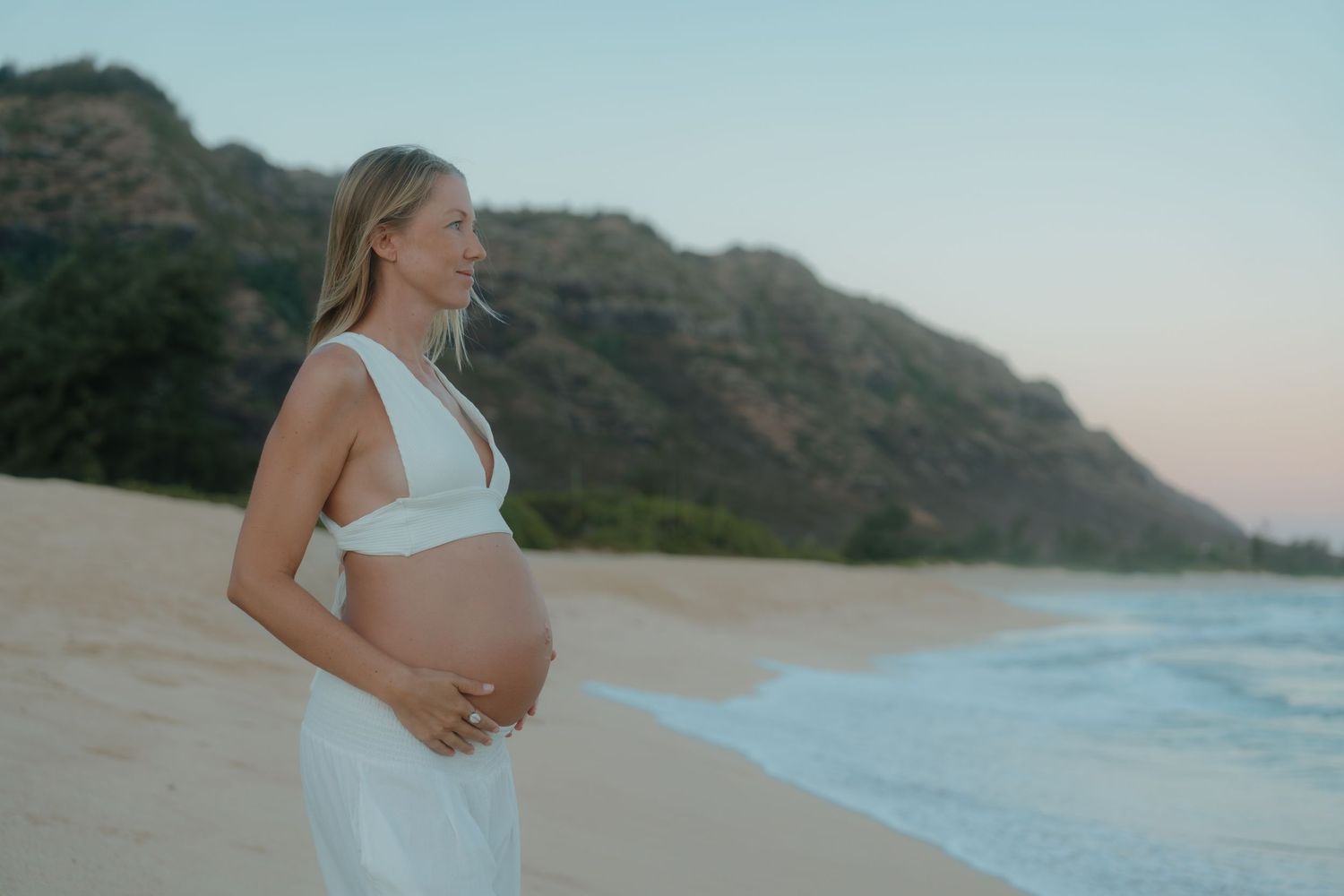 Blonde pregnant woman in white outfit stands on sandy Hawaiian beach at dusk with mountains.