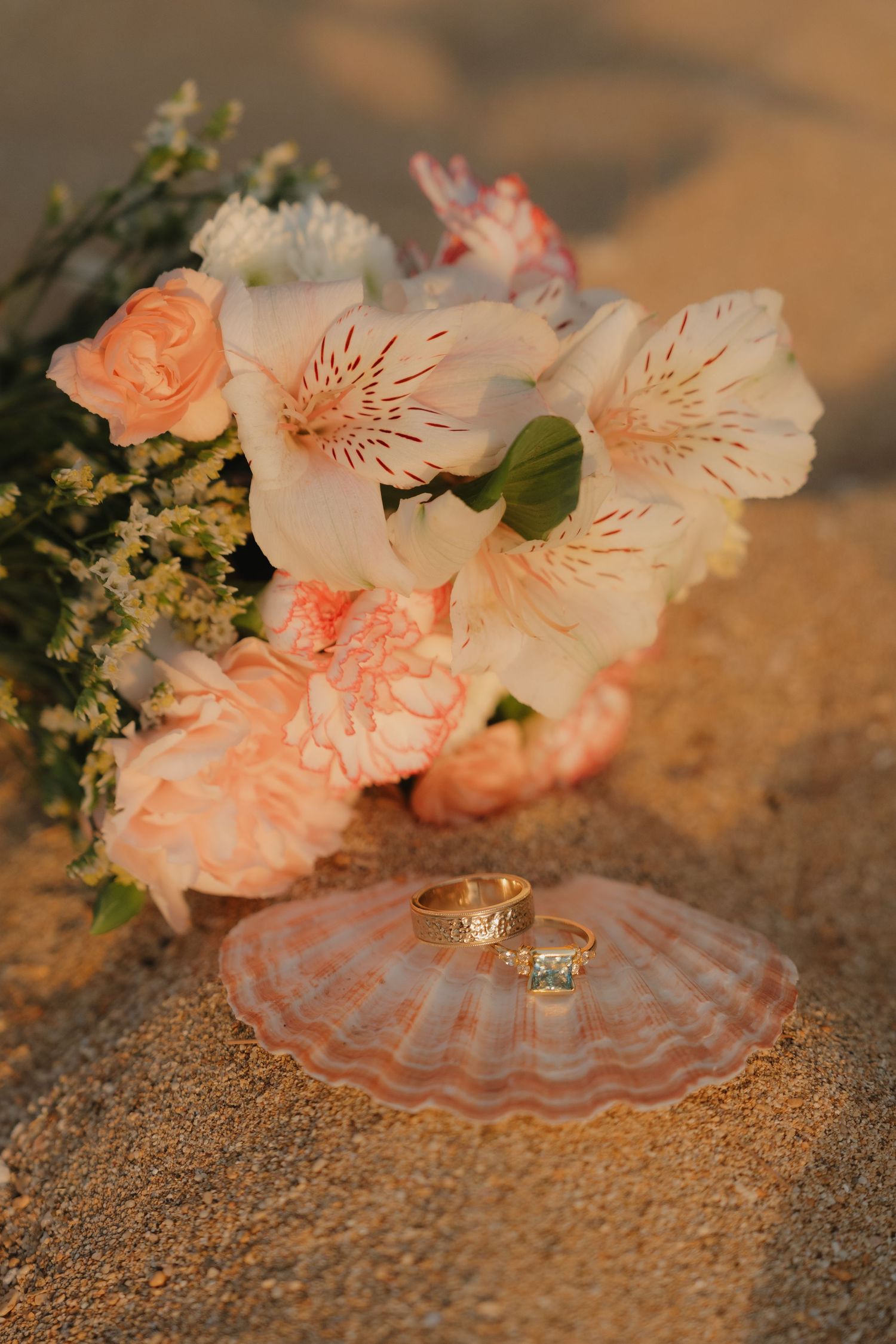Two gold wedding rings rest on a seashell beside a pastel pink bridal bouquet with alstroemeria on sandy beach.
