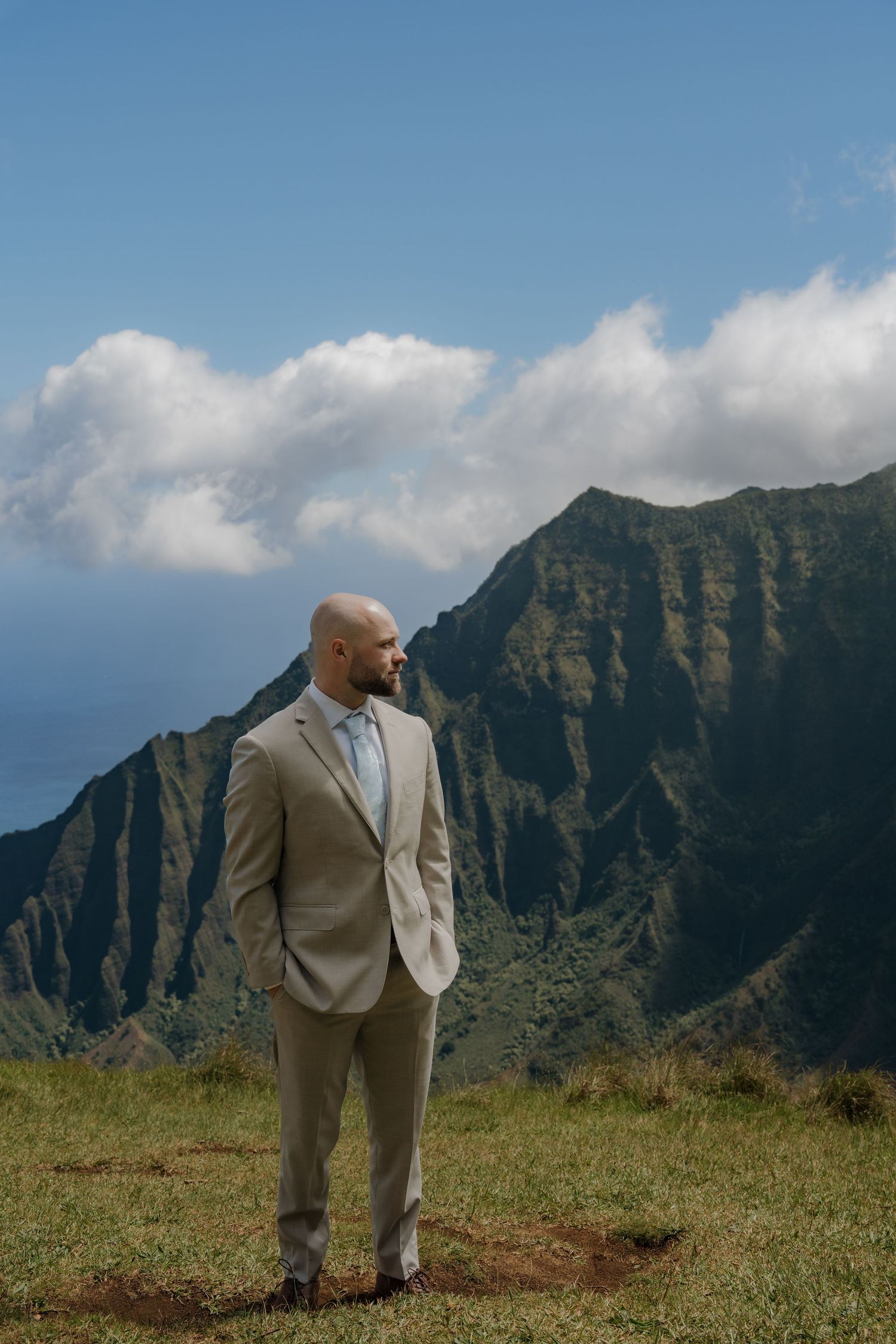 Bald groom in tan suit stands on green hillside with dramatic Na Pali Coast mountains and blue sky in Kauai, Hawaii.