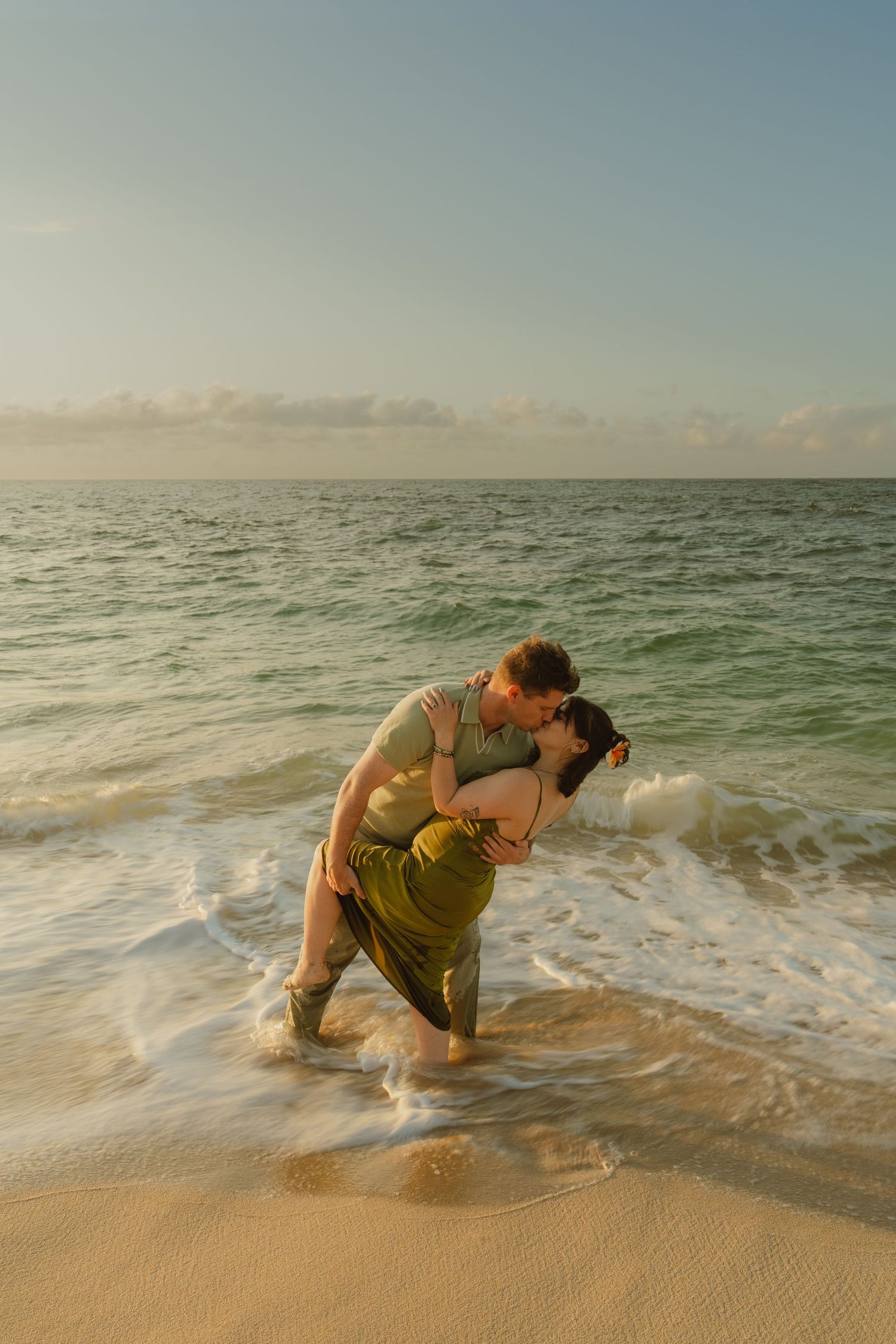 Couple sharing a romantic kiss while standing in ocean waves on a sandy beach at golden hour sunset.