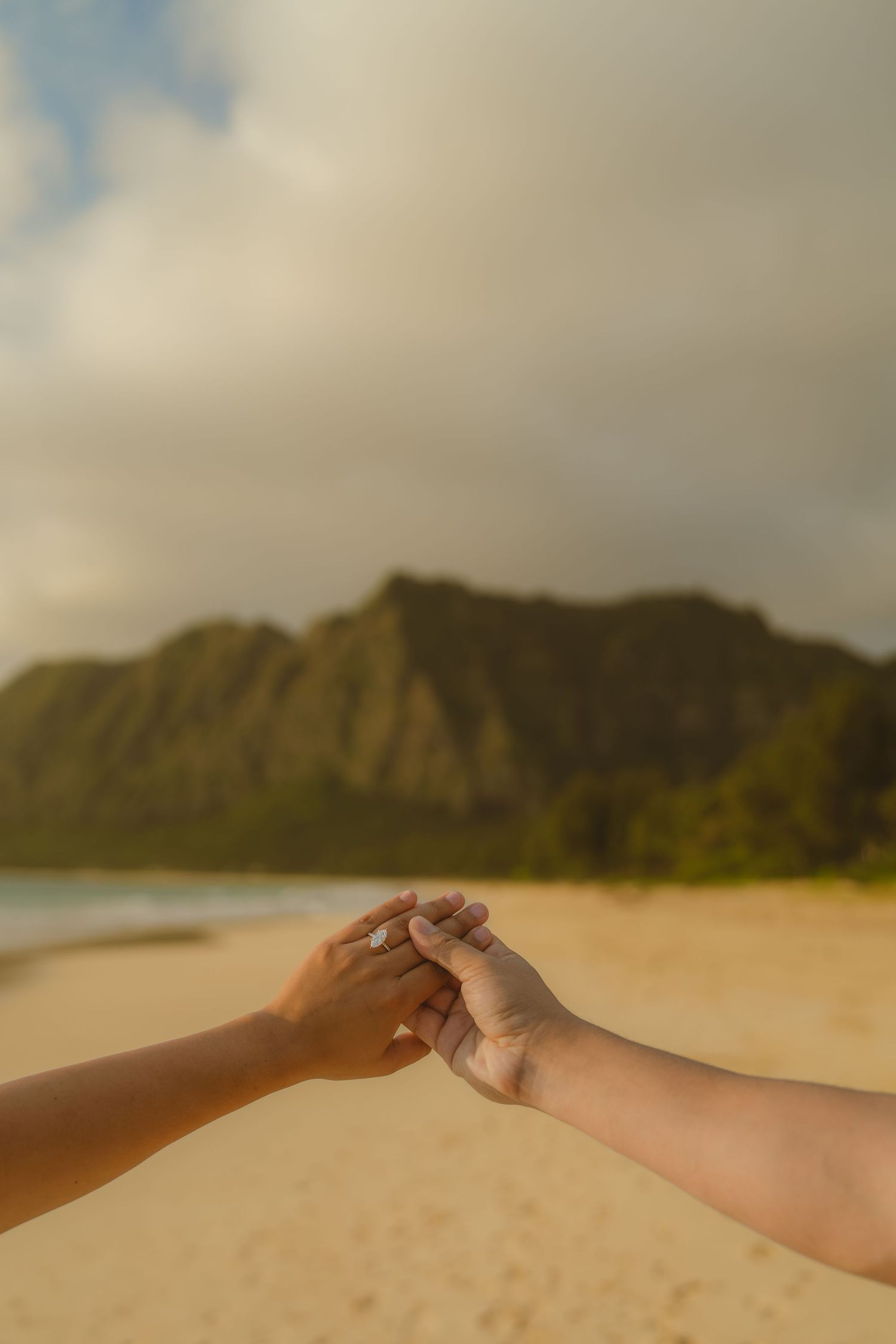 Two people holding hands on a sandy beach with lush green mountains and cloudy sky in Hawaii.