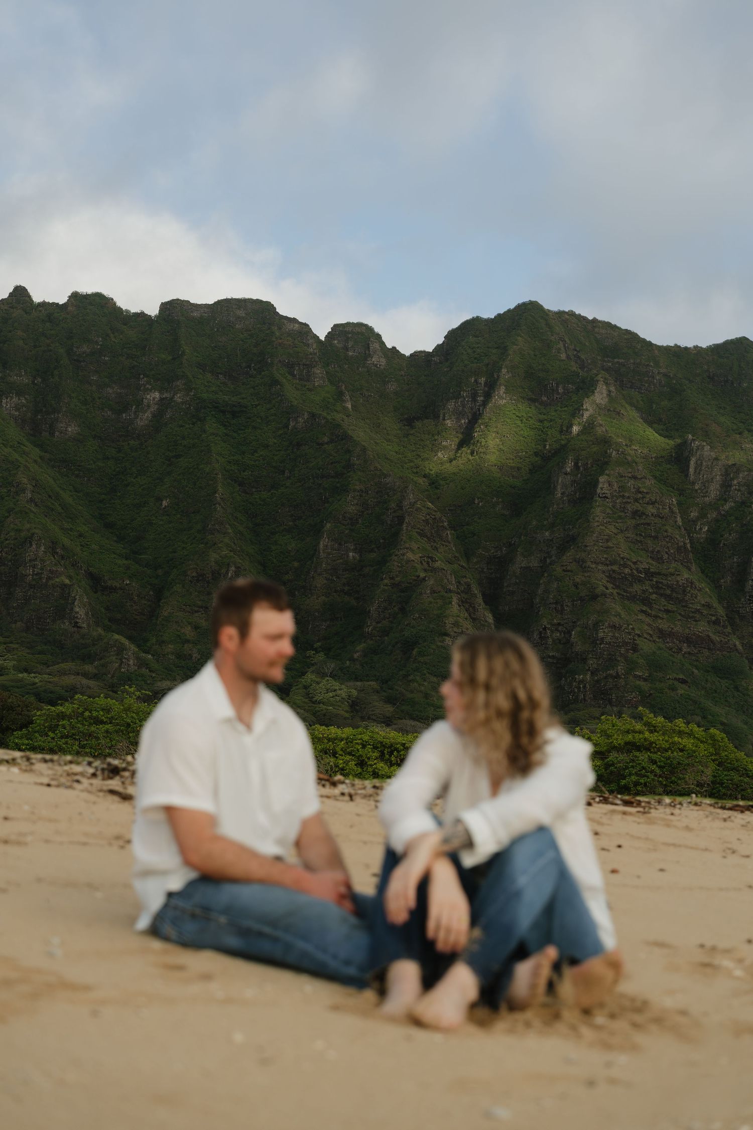 Couple sitting cross-legged on sandy beach with lush green Hawaiian mountains in background.