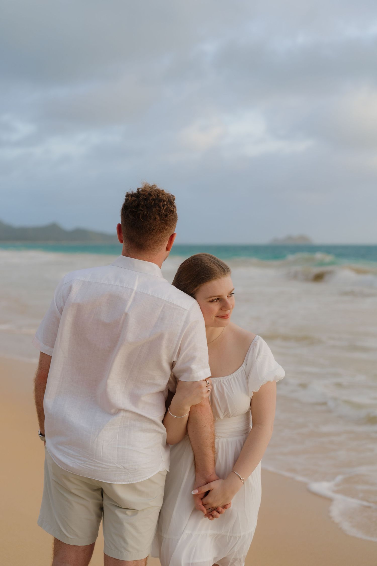 Couple holding hands on a sandy beach, both dressed in white, with ocean waves in the background.