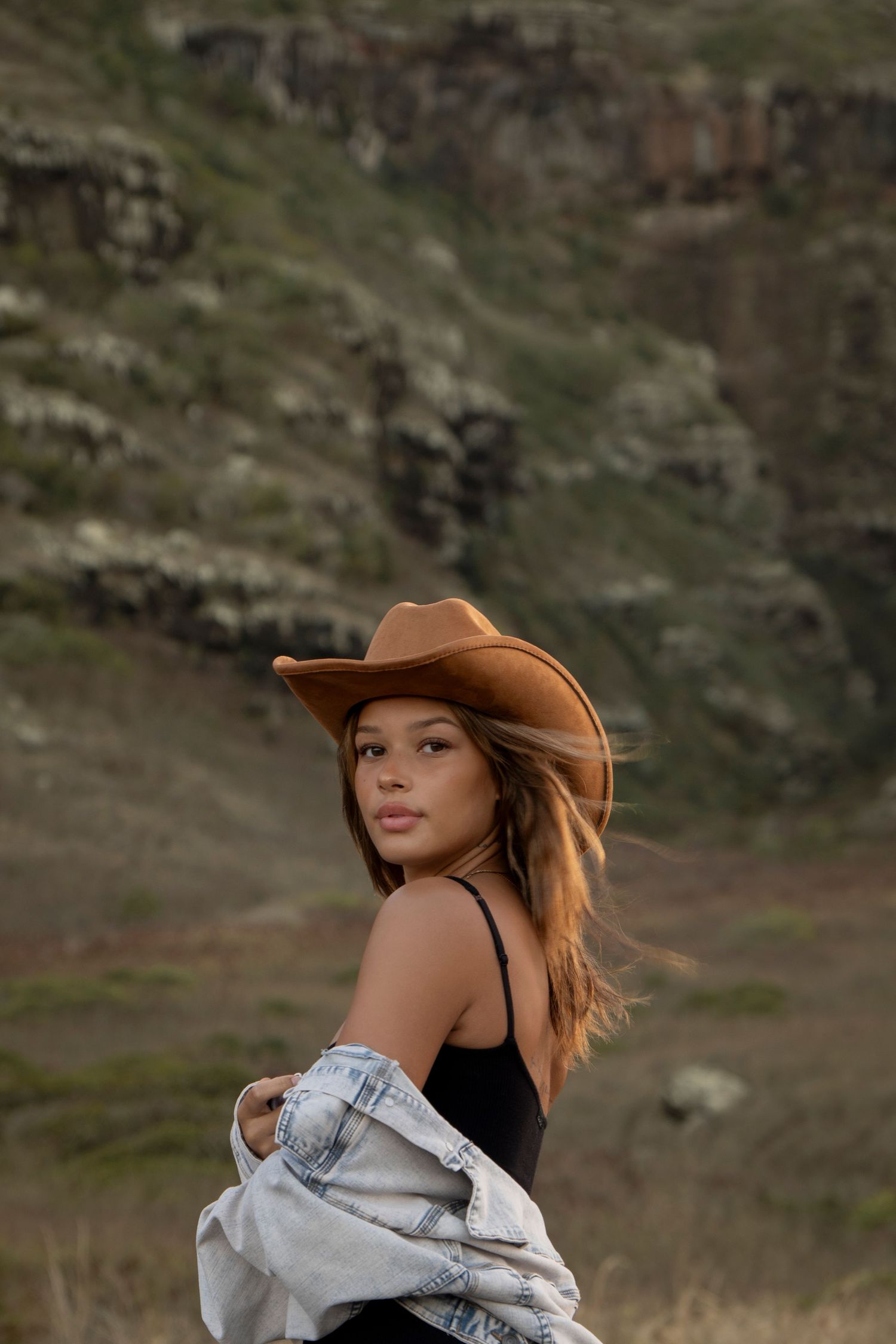 Woman in brown cowboy hat and denim jacket posing against rocky green hillside in golden light.
