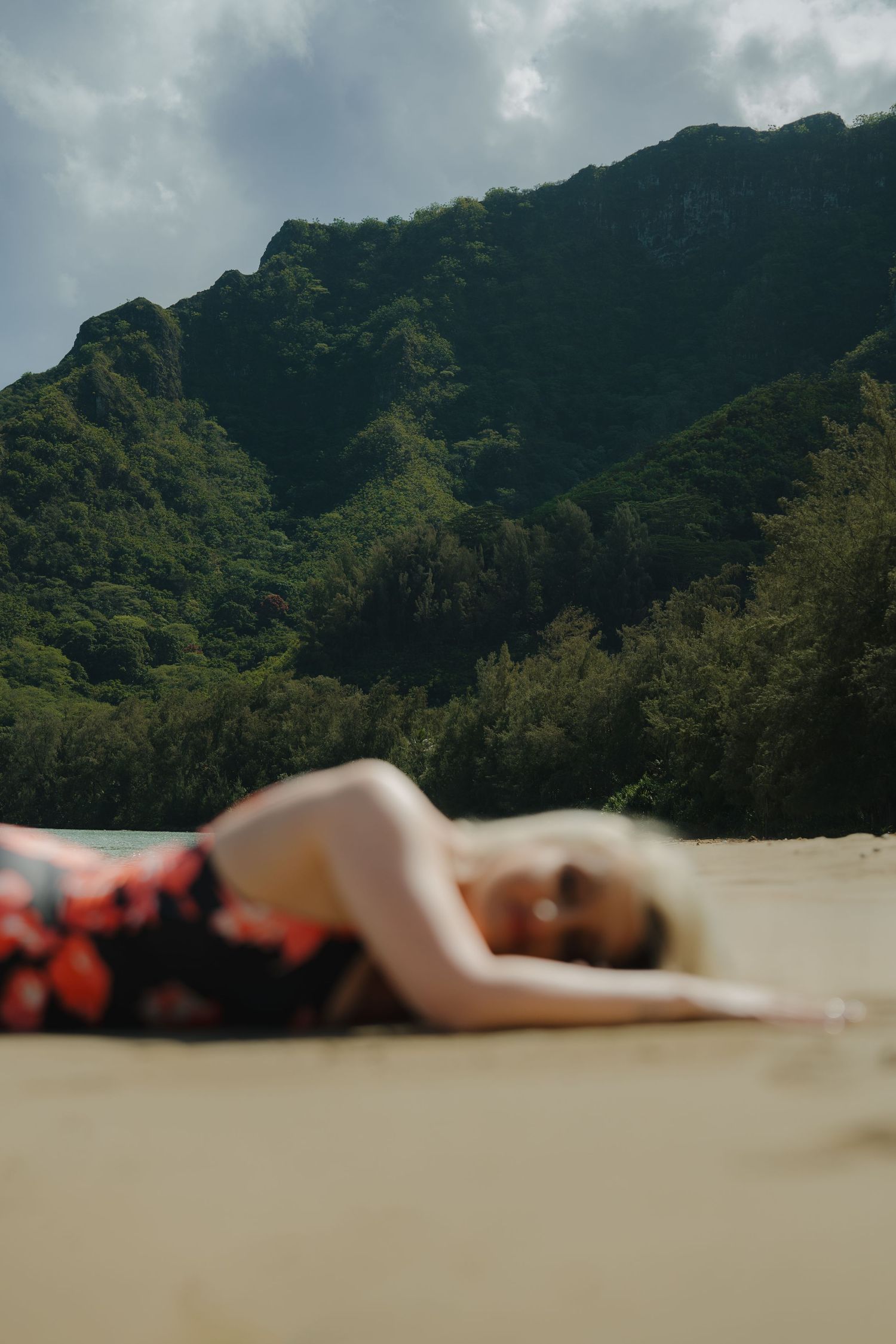 Woman in floral dress lying on sandy beach with dramatic green mountain backdrop.