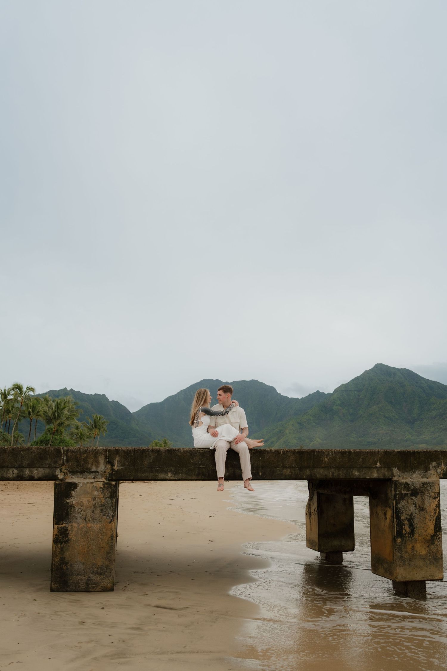 Romantic couple sitting close together on concrete pier at Hanalei Bay beach with mountain backdrop.