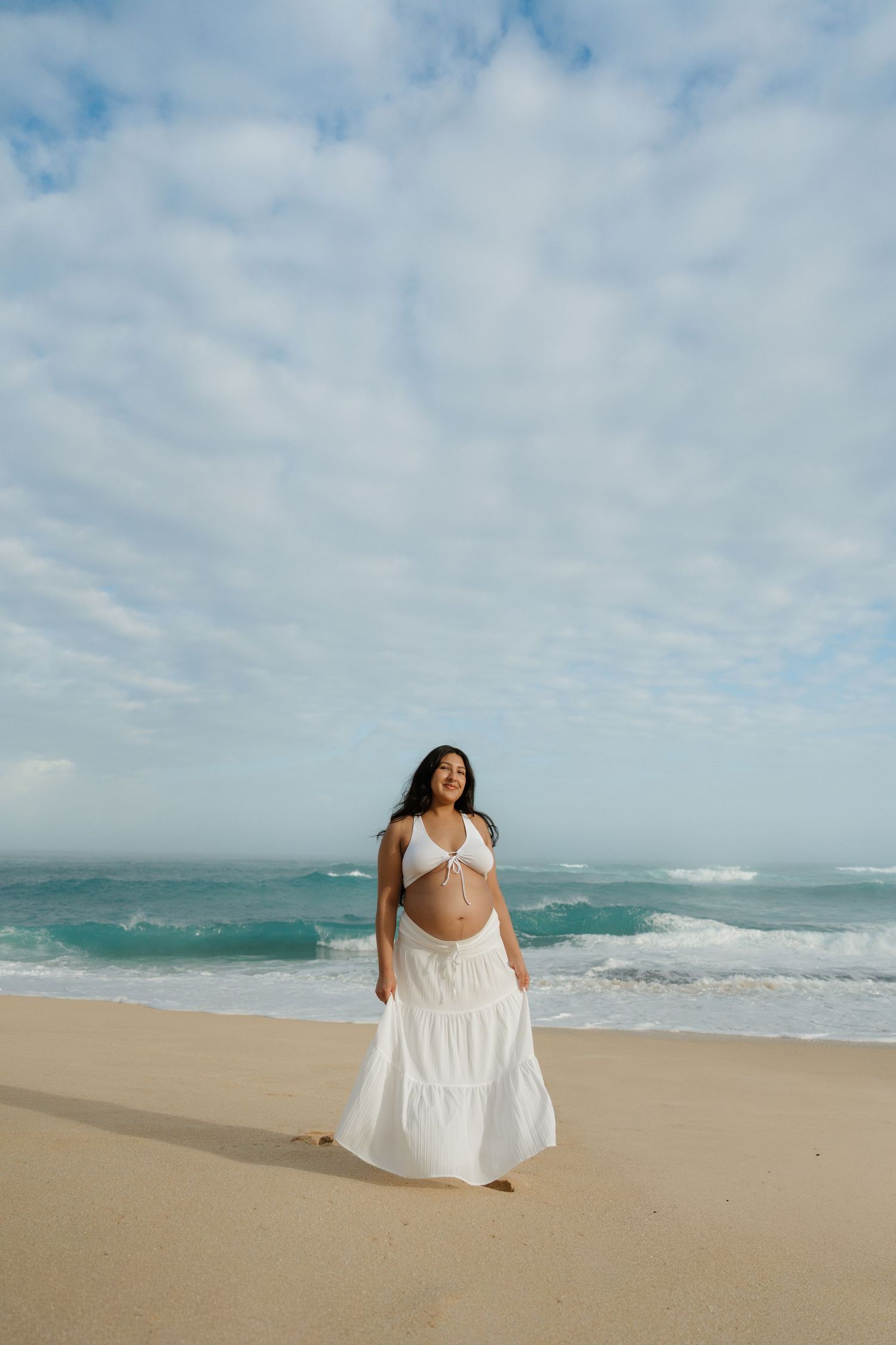Radiant pregnant woman in white two-piece outfit smiling on a sunny beach.