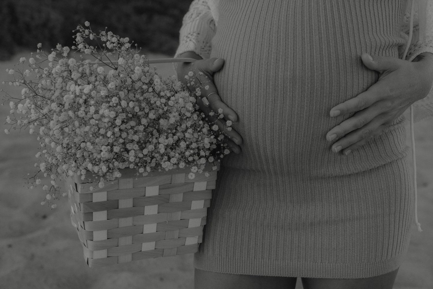 Black and white photo of pregnant woman holding wicker basket of flowers while cradling bump.