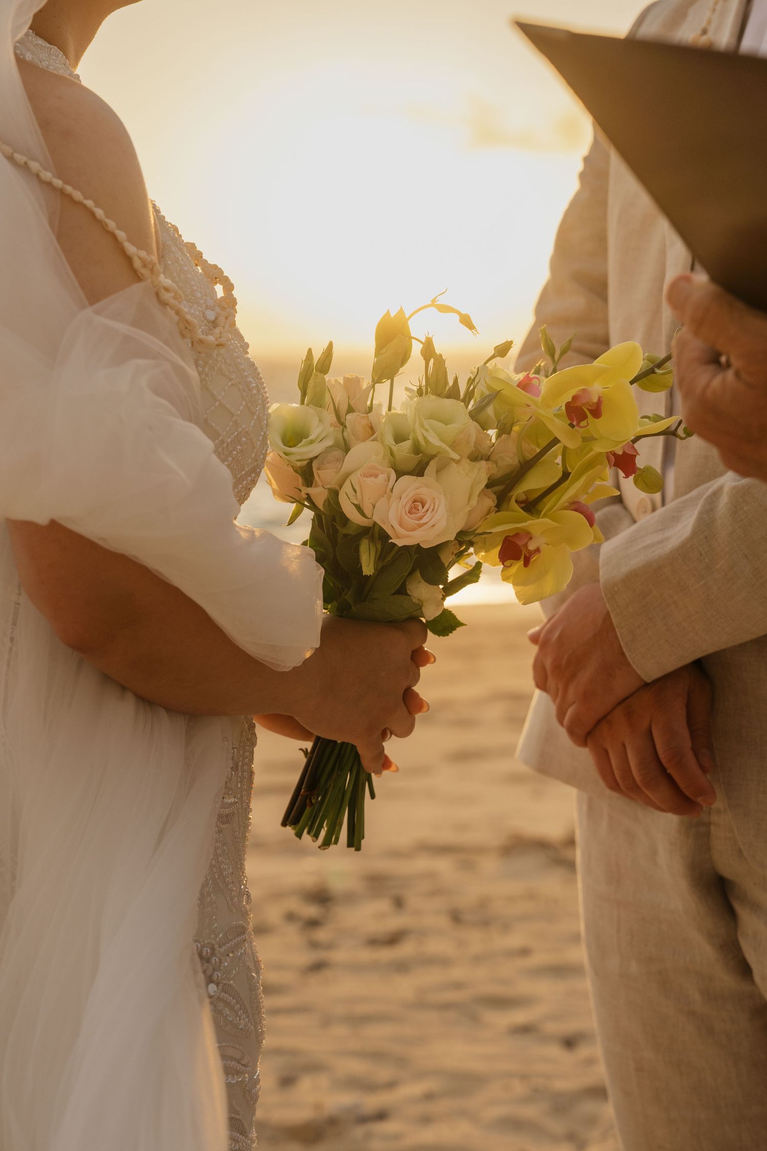 Bride holding a bouquet of white roses and yellow orchids during a beach wedding ceremony at golden sunset.
