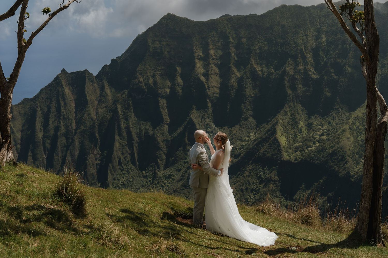 Bride and groom embrace on lush Hawaiian hillside with dramatic Na Pali Coast mountain cliffs backdrop.