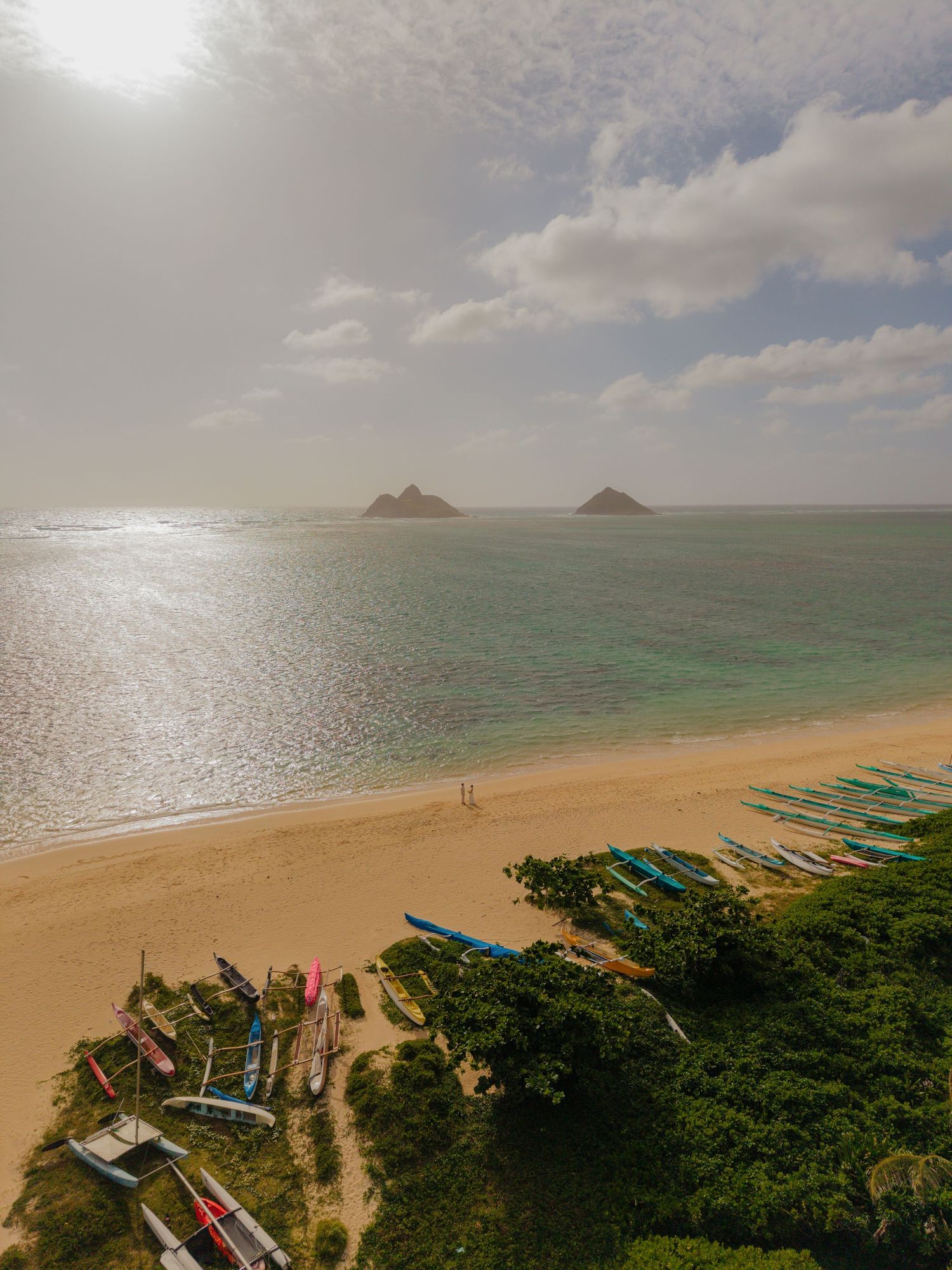 Aerial view of Lanikai Beach Hawaii with colorful outrigger canoes on golden sand and twin Mokulua Islands in turquoise water.