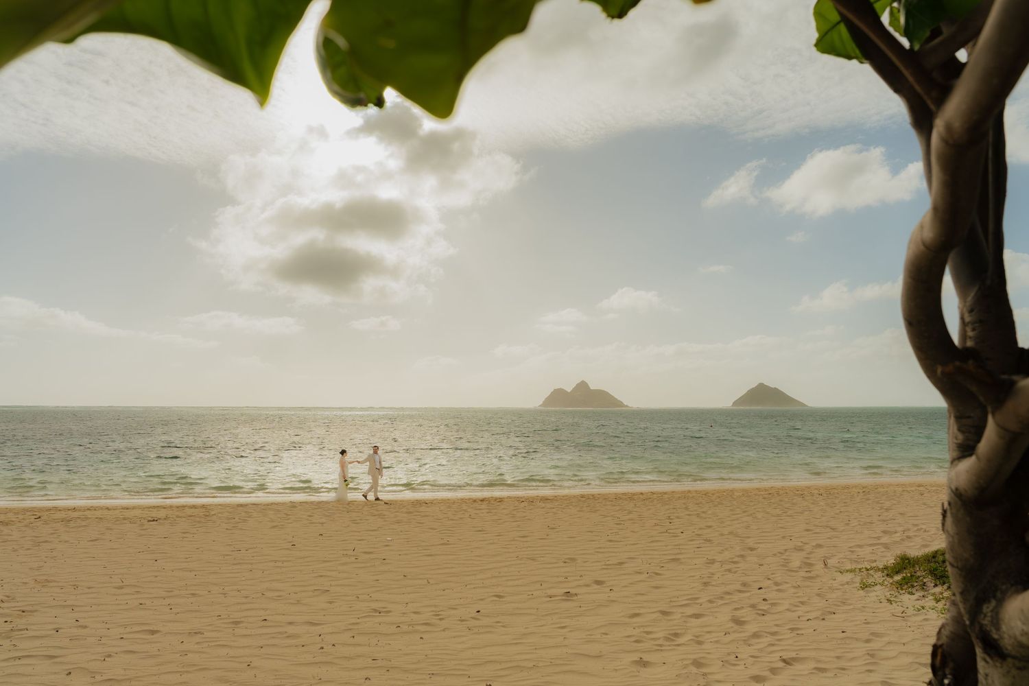 Couple walking on a sandy Hawaiian beach with turquoise ocean waters and Mokulua Islands visible in the background.
