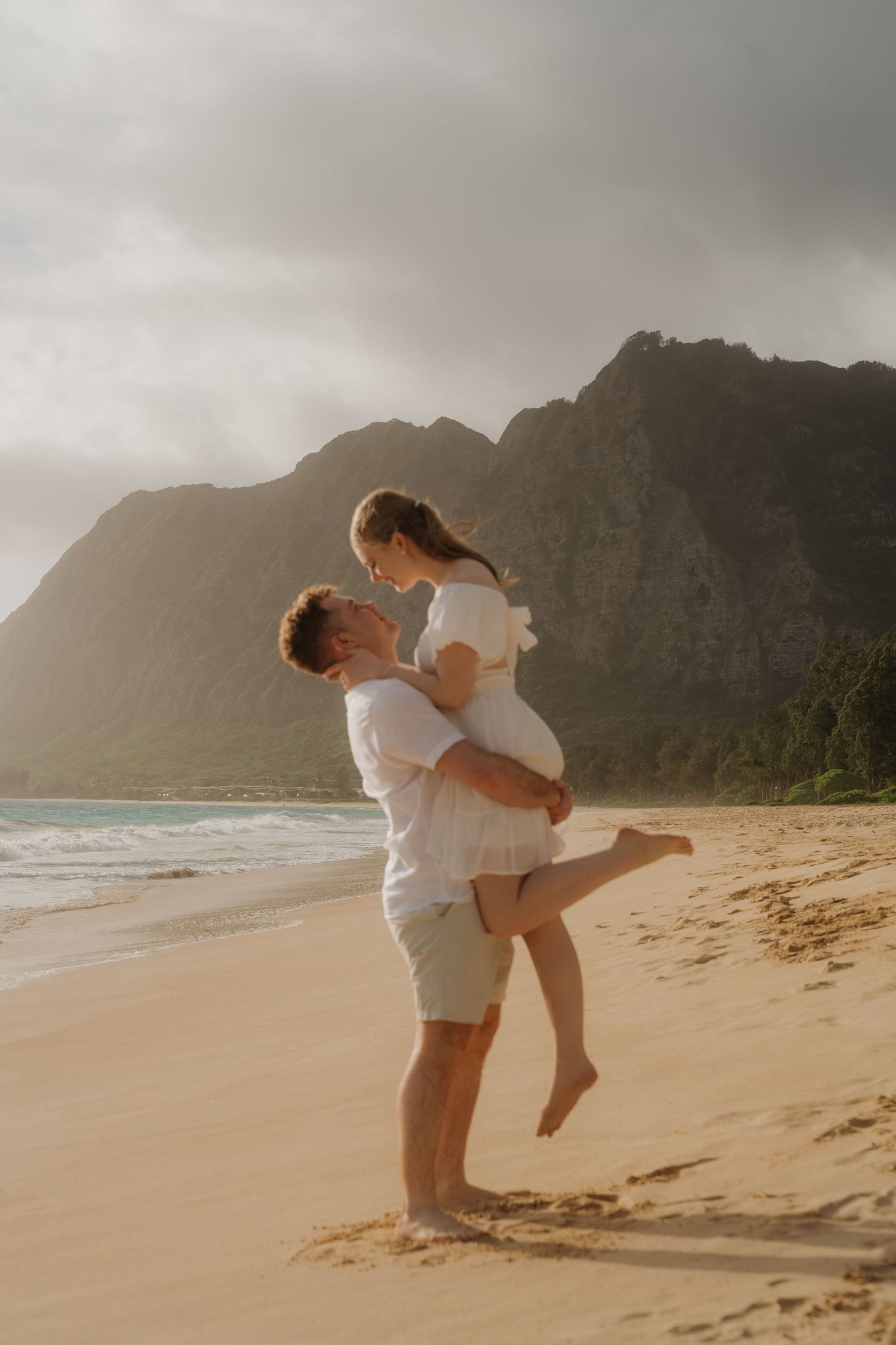 Man lifting woman on a sandy beach with dramatic mountain backdrop at golden hour sunset.