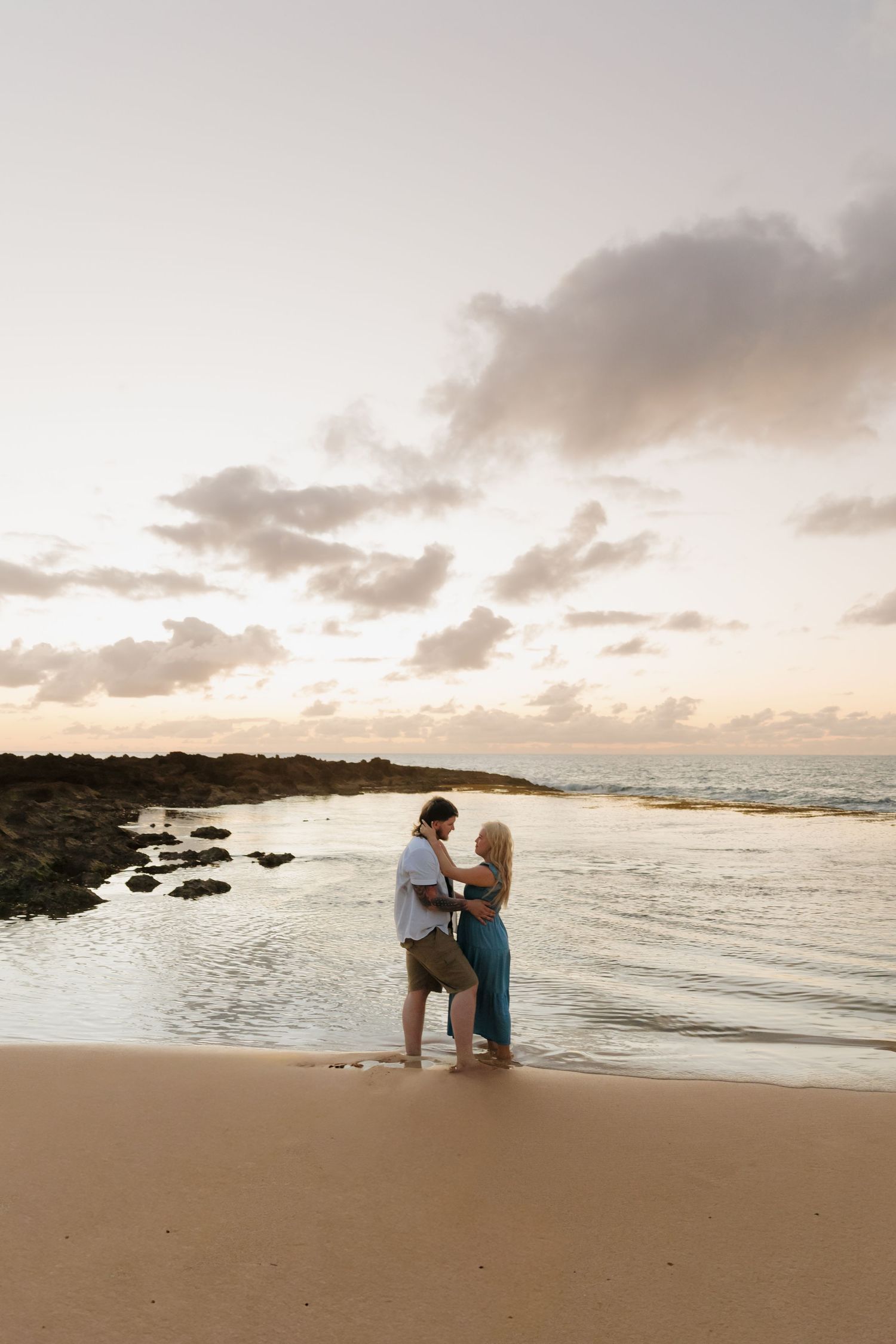 Couple embracing on a sandy beach at sunset with calm ocean waves and rocky shoreline in the background.