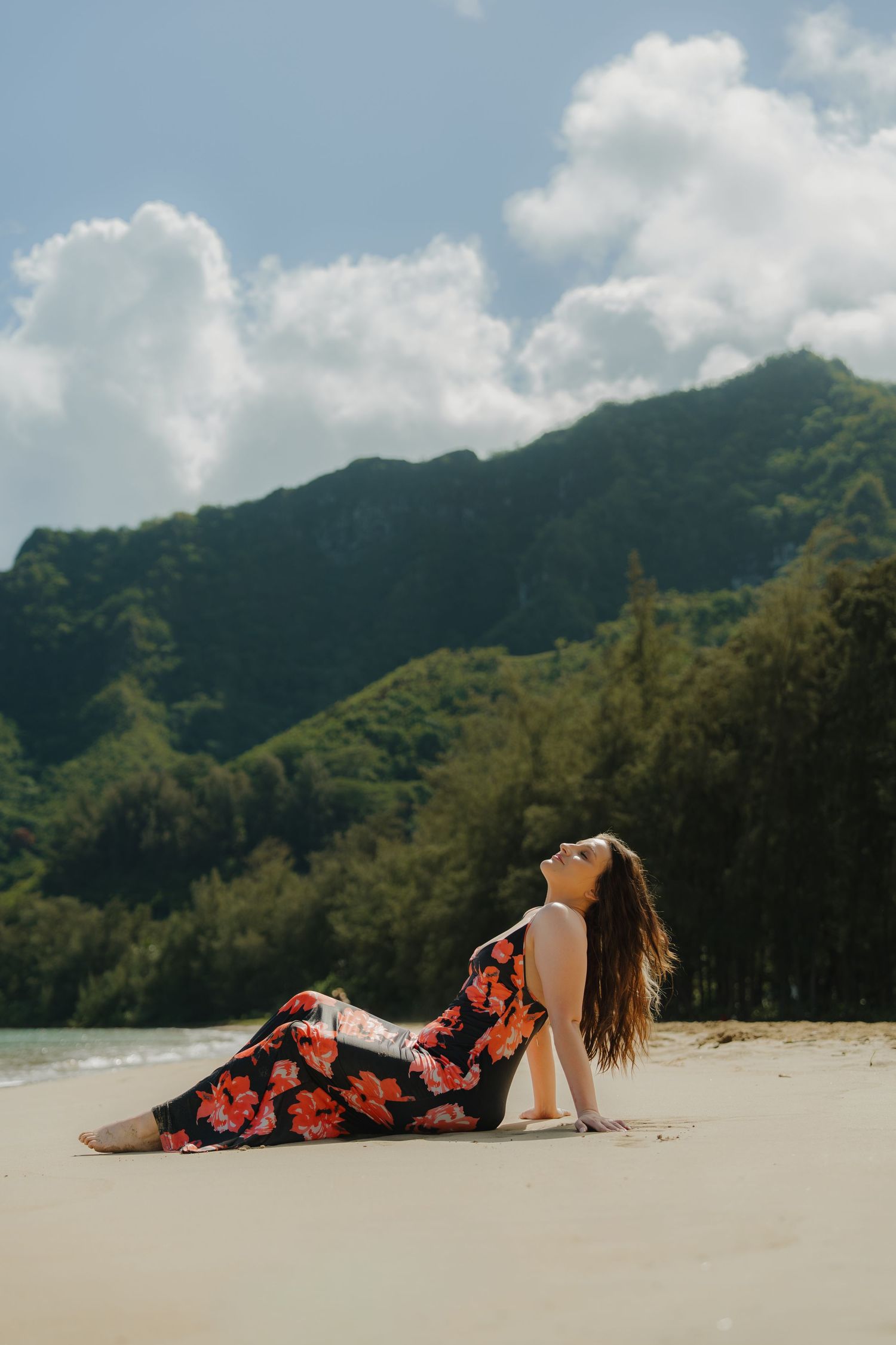Woman in floral dress reclining on sandy beach with lush green mountains behind.