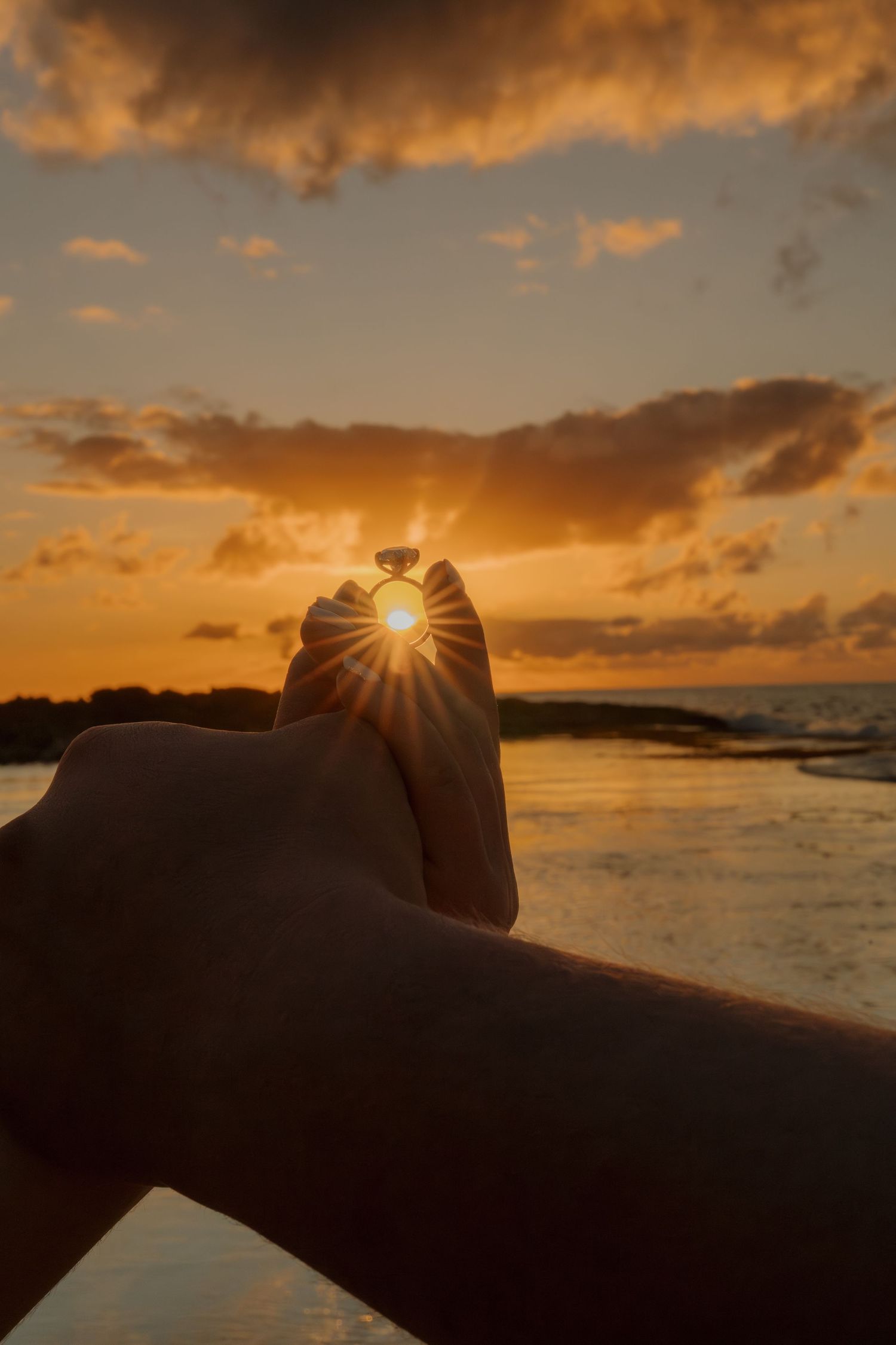 Hand holding a ring up to a golden sunset over the ocean, with sun rays bursting through the diamond.