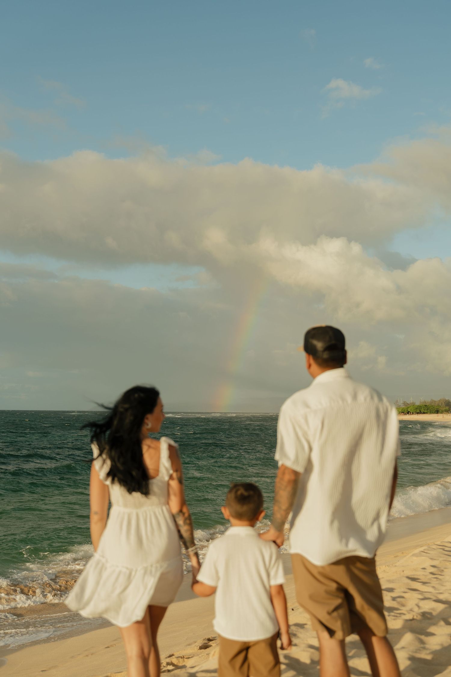 Family of three holding hands on a sandy beach watching a rainbow over the ocean.