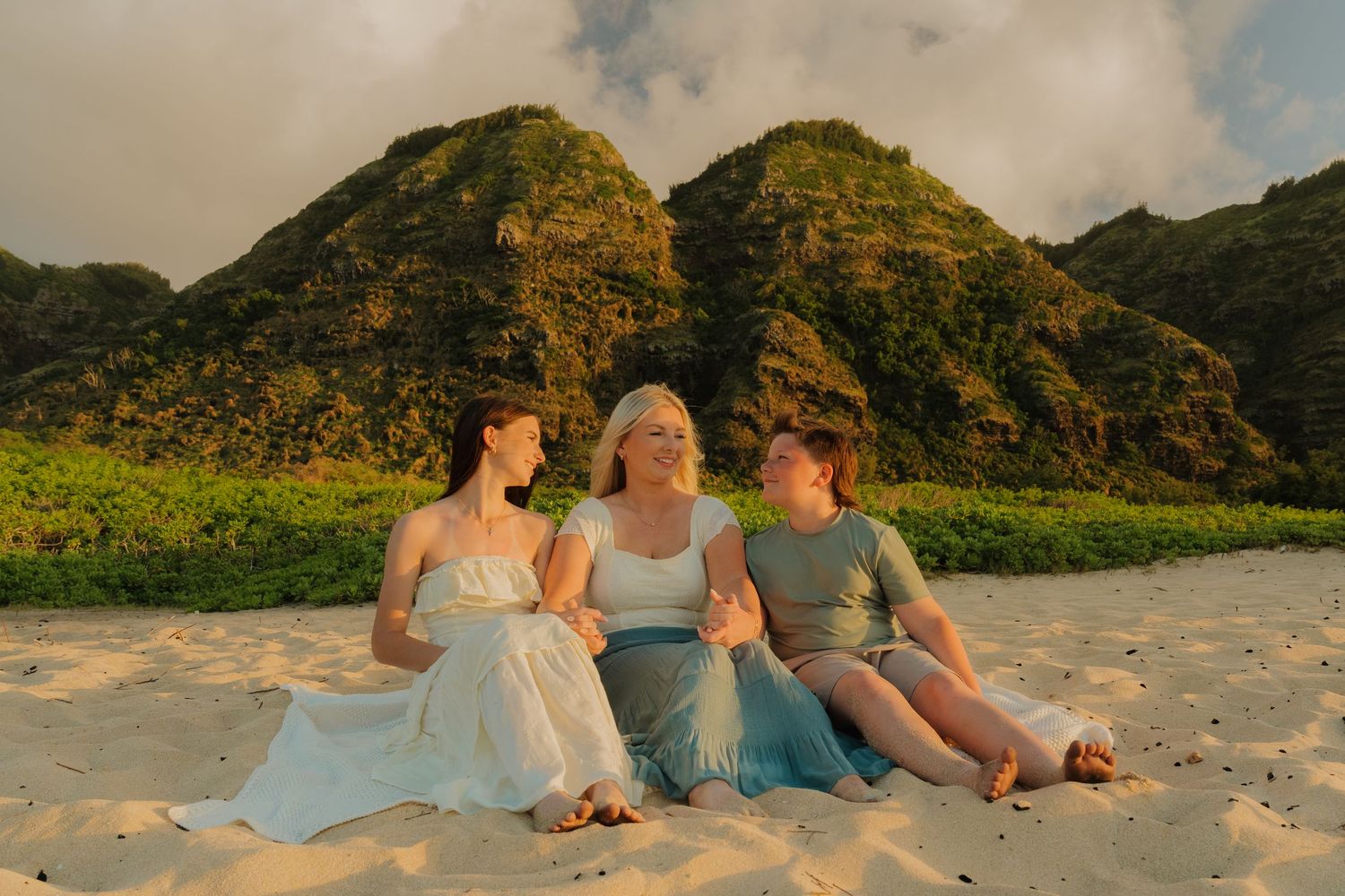 Three women sitting together on a sandy beach with lush green mountains at golden hour in Hawaii.