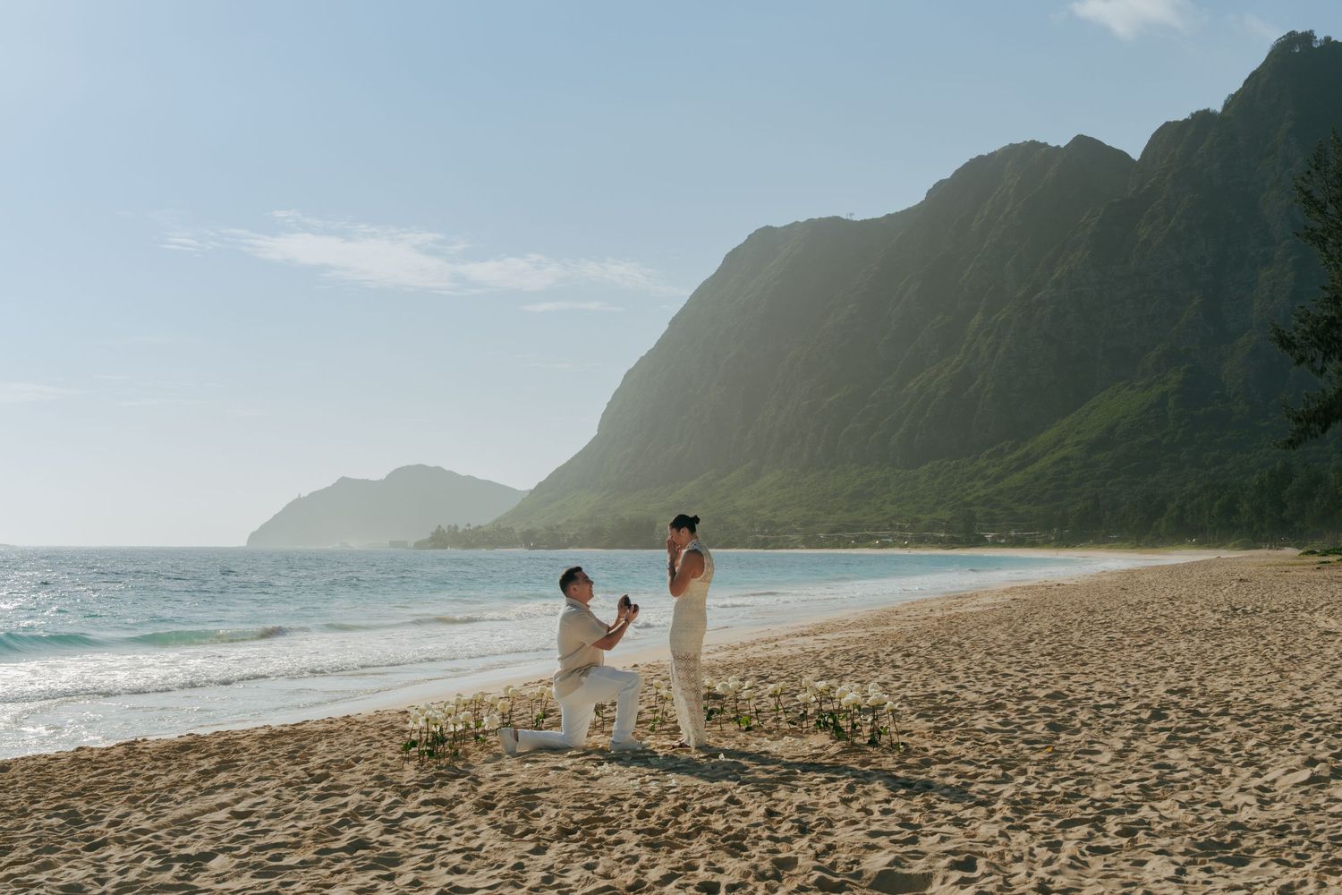 A couple shares a romantic beach proposal moment on a sandy Hawaiian shore with lush green mountains in the background.