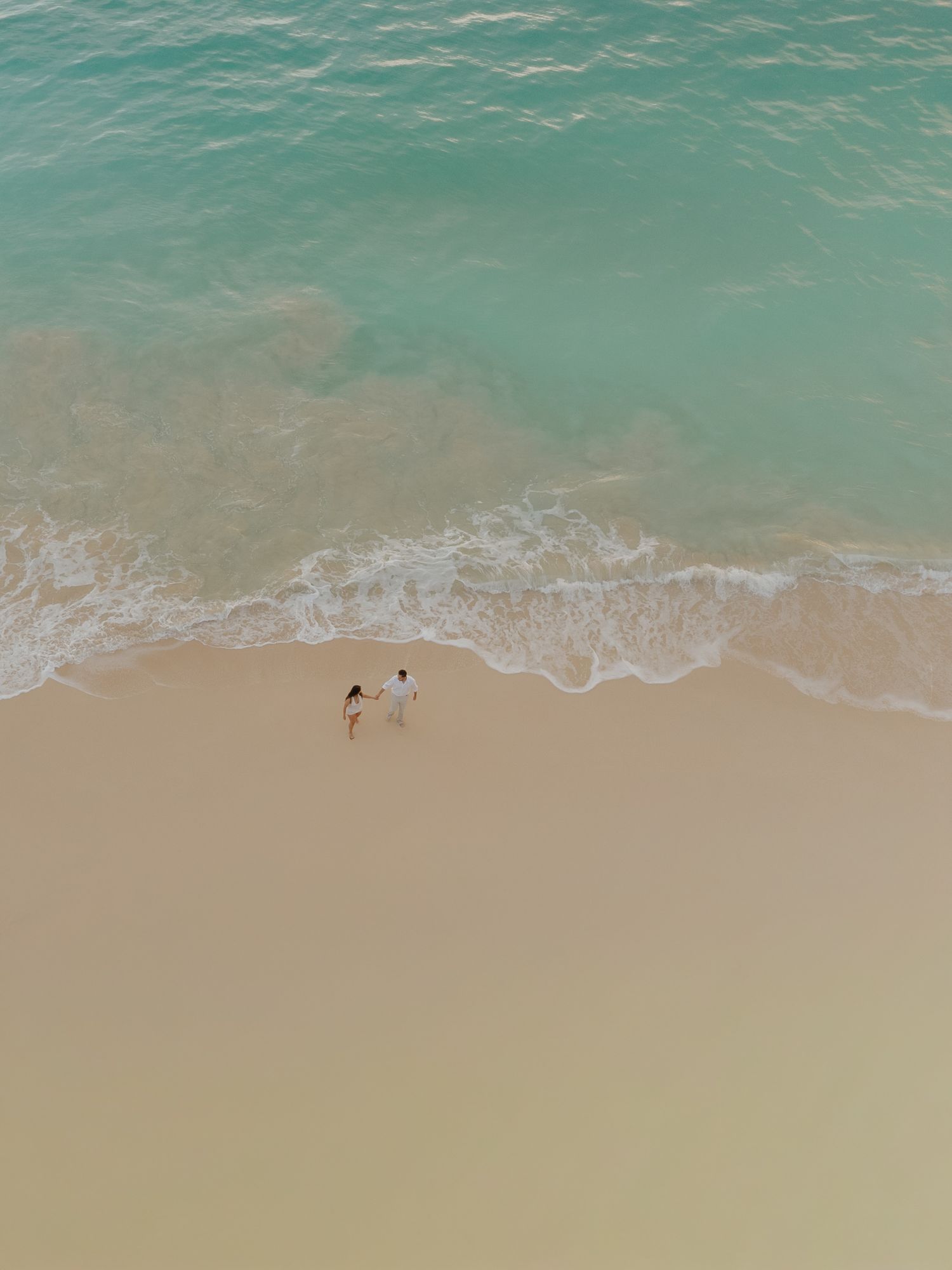 Aerial drone view of couple holding hands walking along turquoise shoreline.