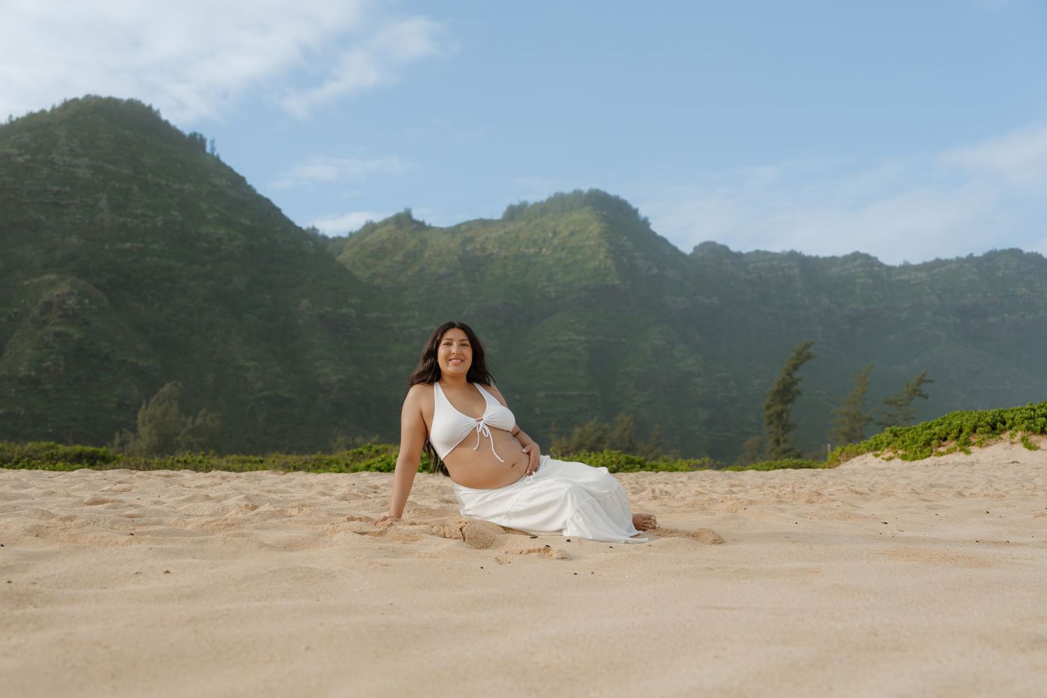 Pregnant woman in white outfit sitting on sandy beach with lush green mountains backdrop in Hawaii.