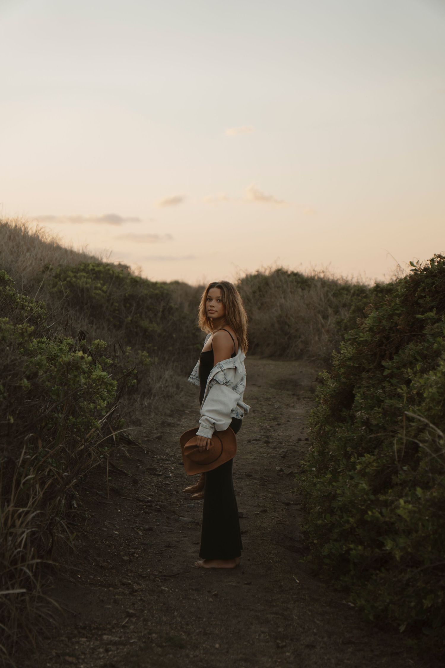 Woman in denim jacket holding cowboy hat standing on dirt path surrounded by coastal shrubs at dusk.