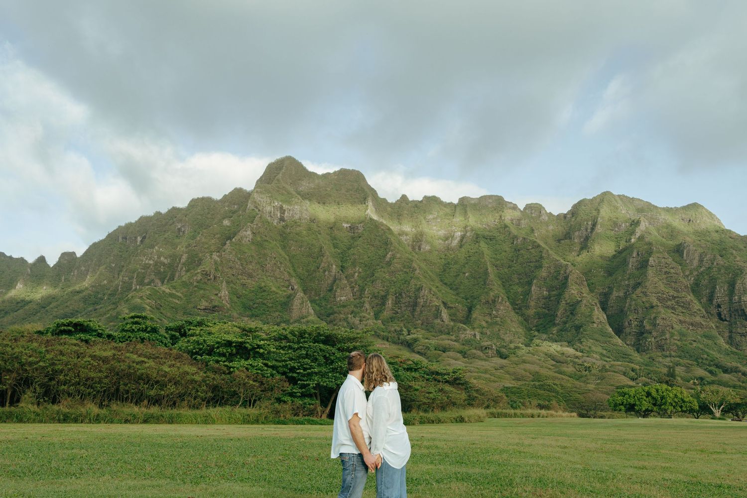 Couple holding hands on green field with dramatic jagged Hawaiian mountain range behind them.