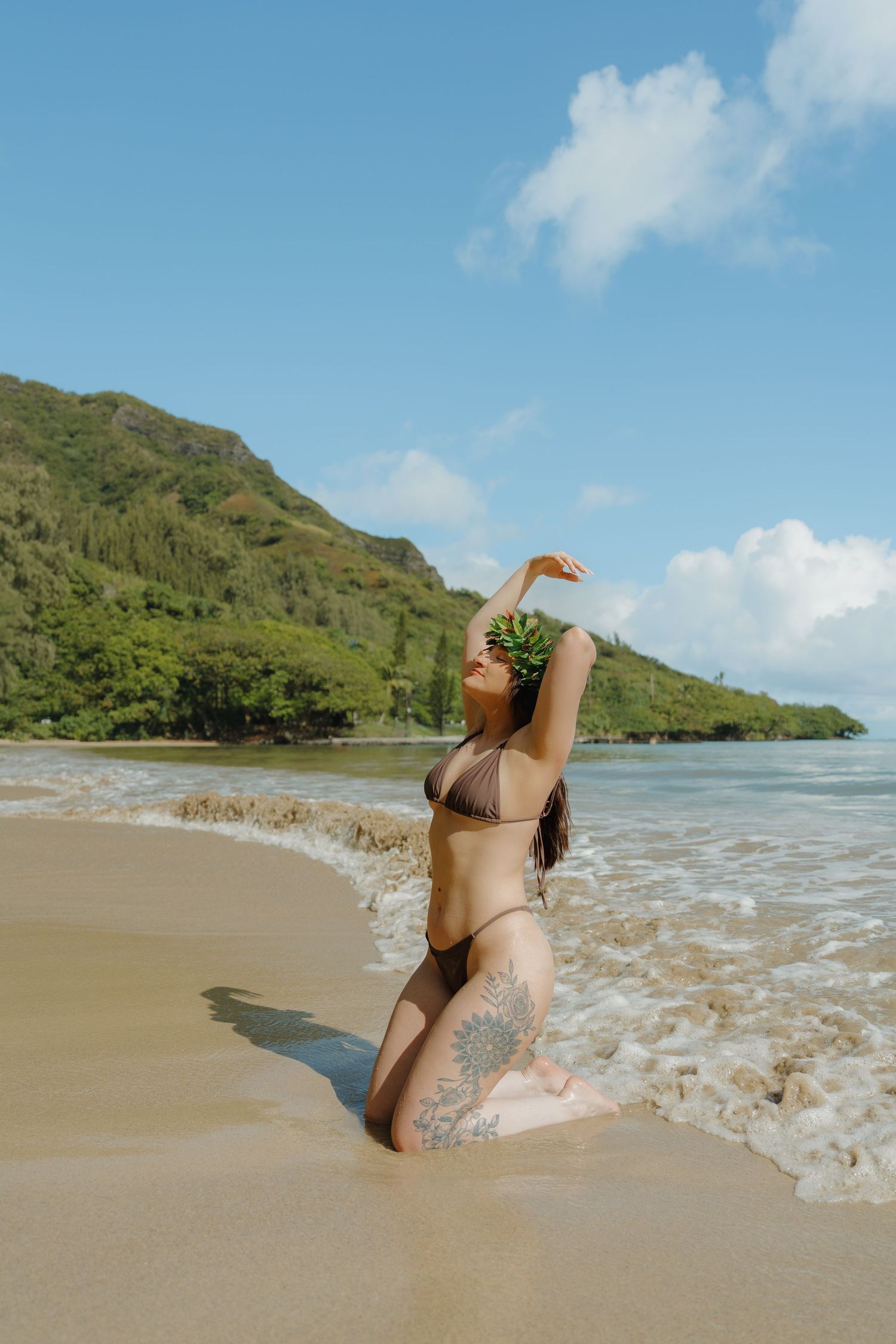 Tattooed woman in brown bikini with leaf crown posing in ocean waves on tropical beach.