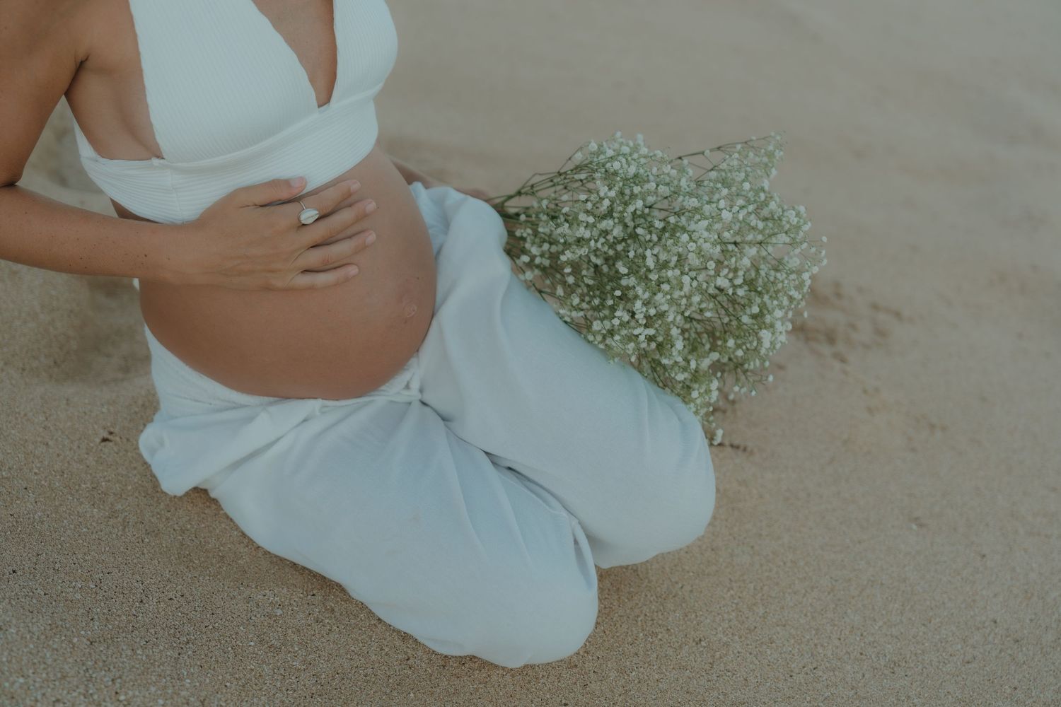 Pregnant woman in white sitting on sand holding baby bump with floral bouquet beside her.
