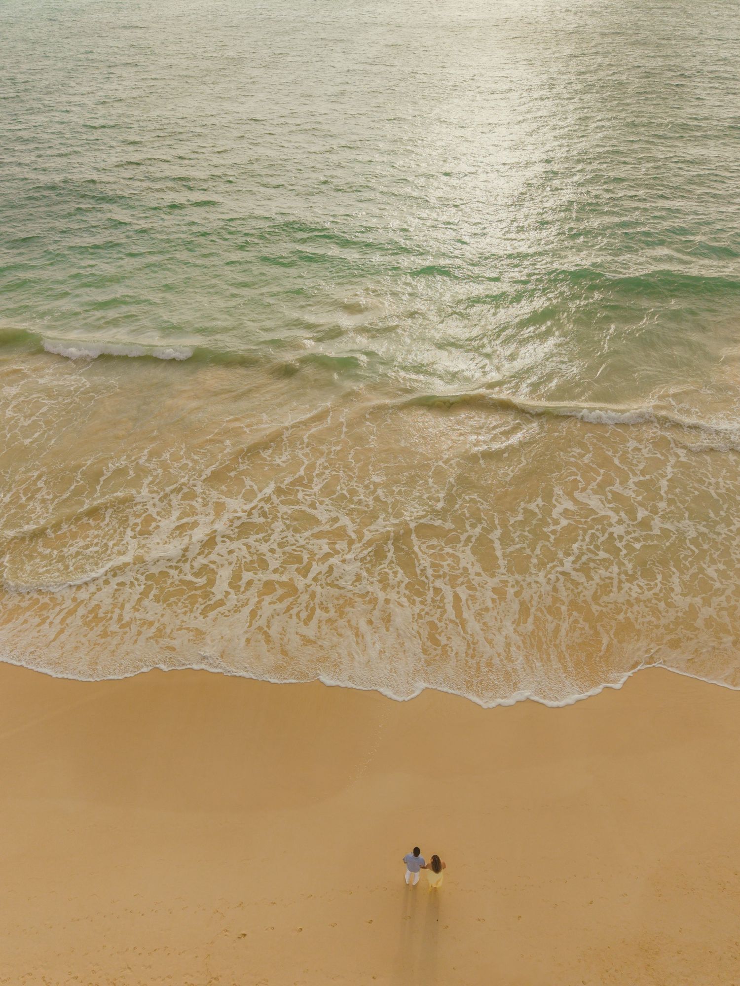 Aerial view of two people standing on a sandy beach where gentle turquoise waves meet the shore.