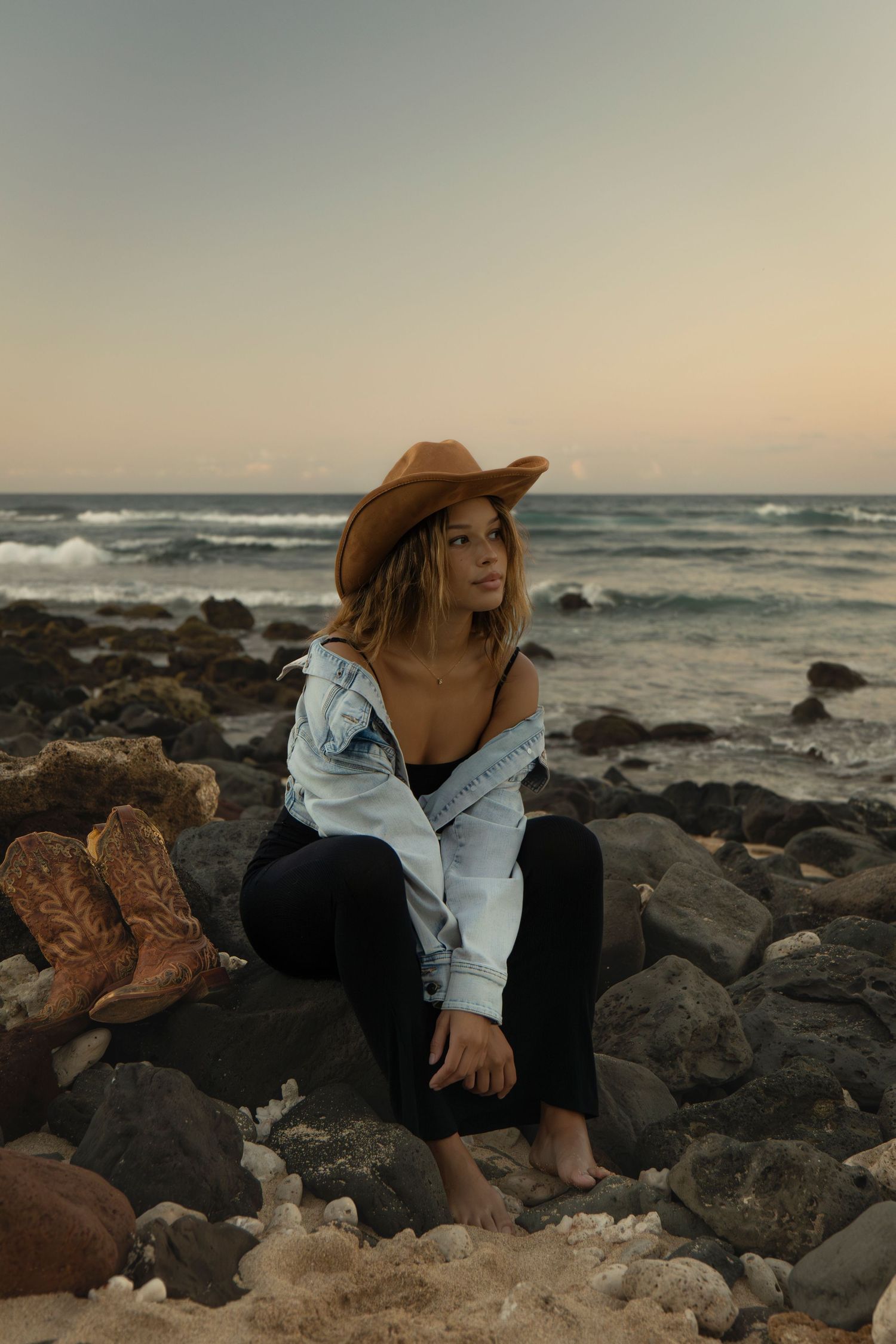 Woman in cowboy hat and denim jacket sitting on rocky beach at sunset with boots nearby.