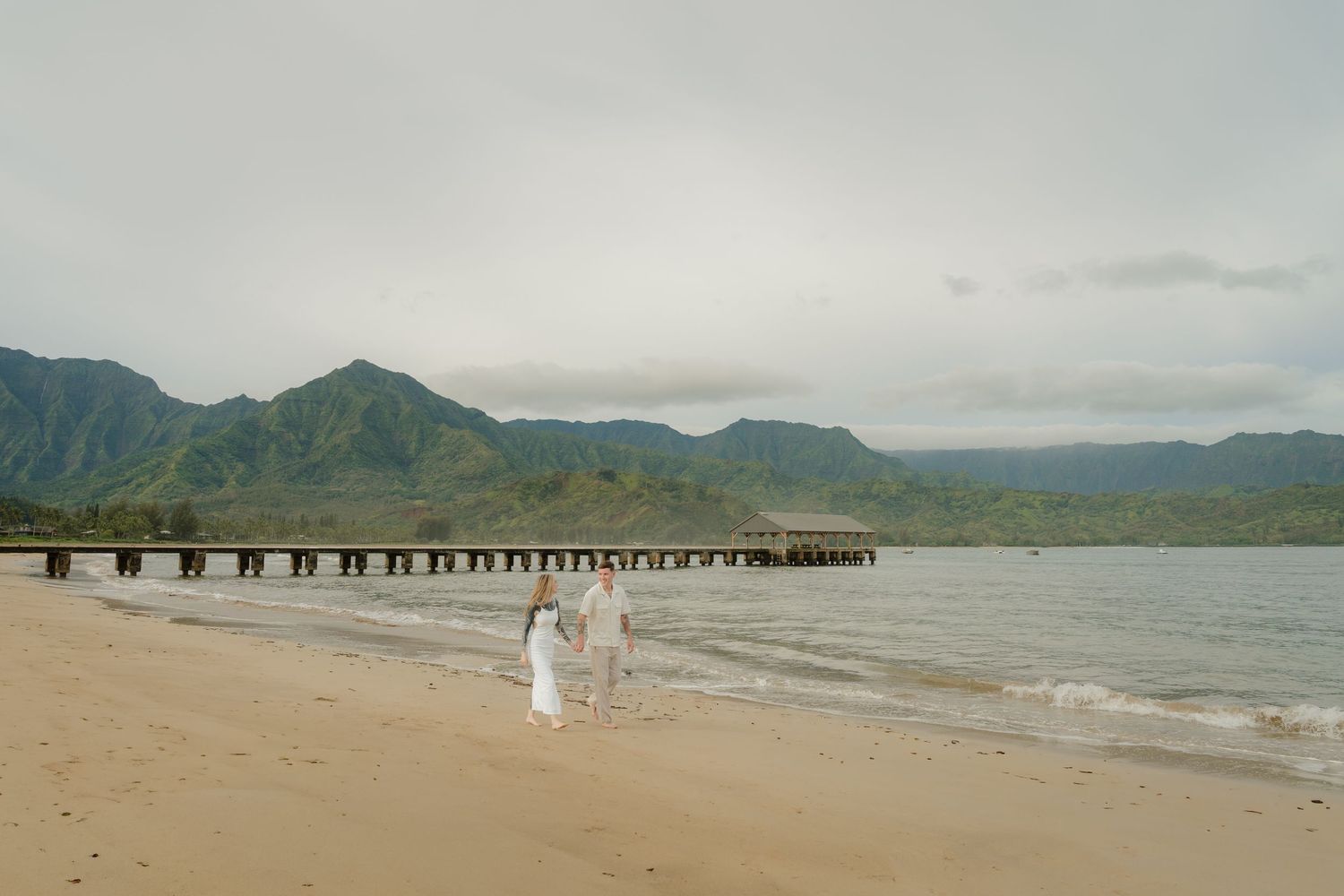 Couple walking hand in hand along Hanalei Bay shoreline with iconic pier and green mountains.
