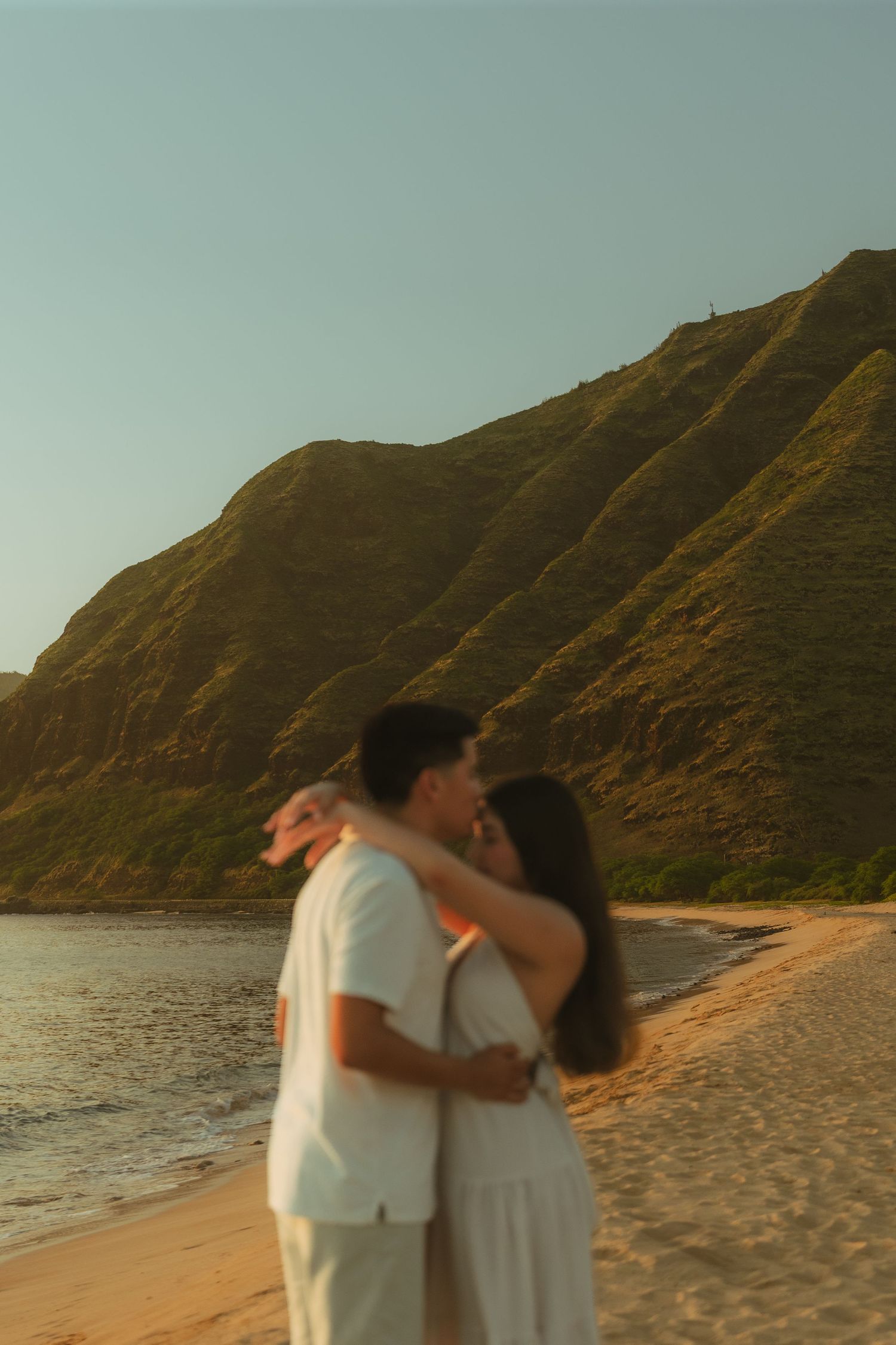 A couple shares a romantic kiss on a sandy Hawaiian beach at golden hour with lush green mountains in the background.
