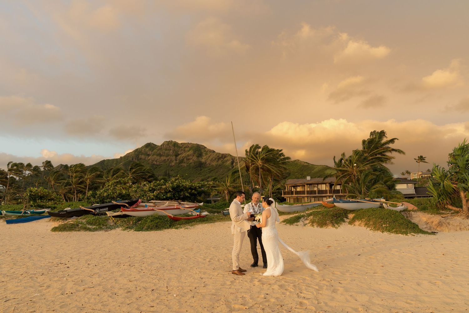 Couple exchanges vows on a sandy Hawaiian beach at golden sunset with lush mountains and palm trees in the background.