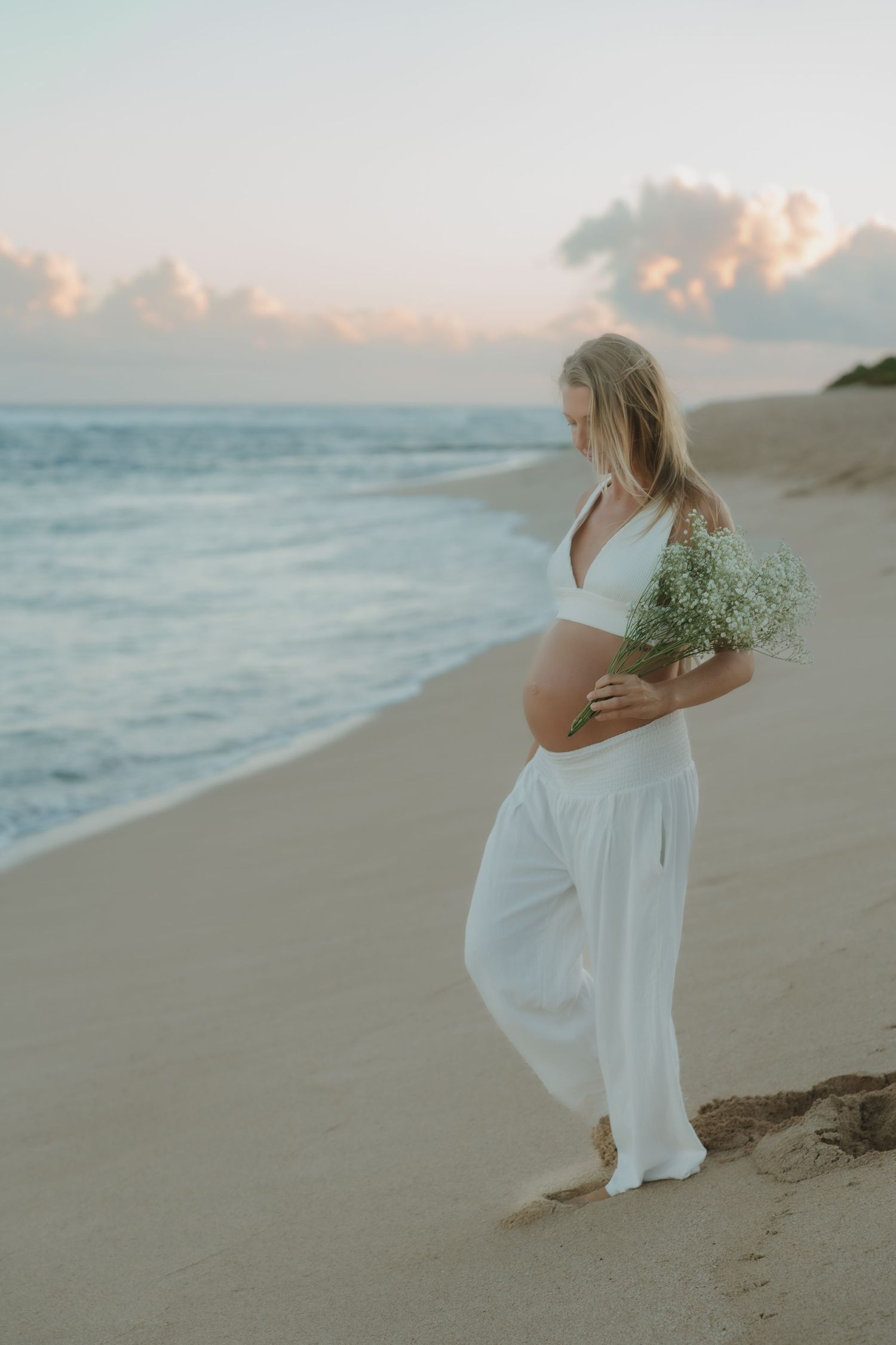 Pregnant woman in white holding baby's breath bouquet on sandy beach at sunset.