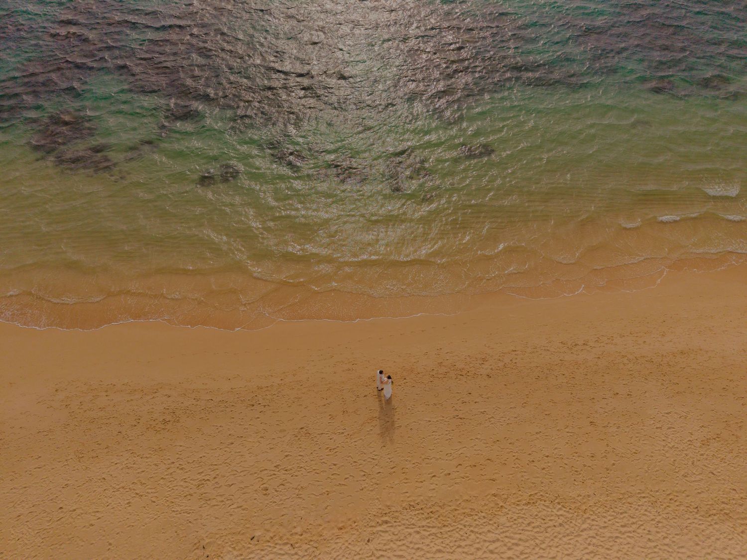 Aerial view of two people standing on a sandy tropical beach beside clear turquoise ocean waters.