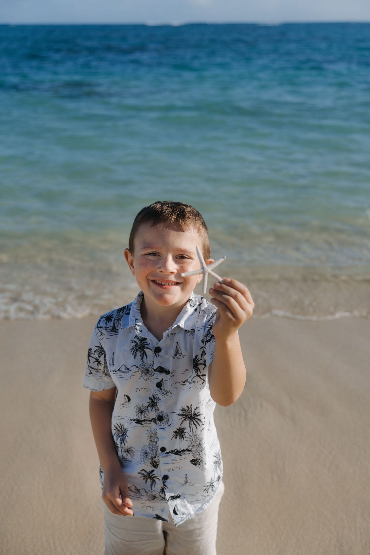 Happy young boy smiling and holding a small starfish on a sunny tropical beach.
