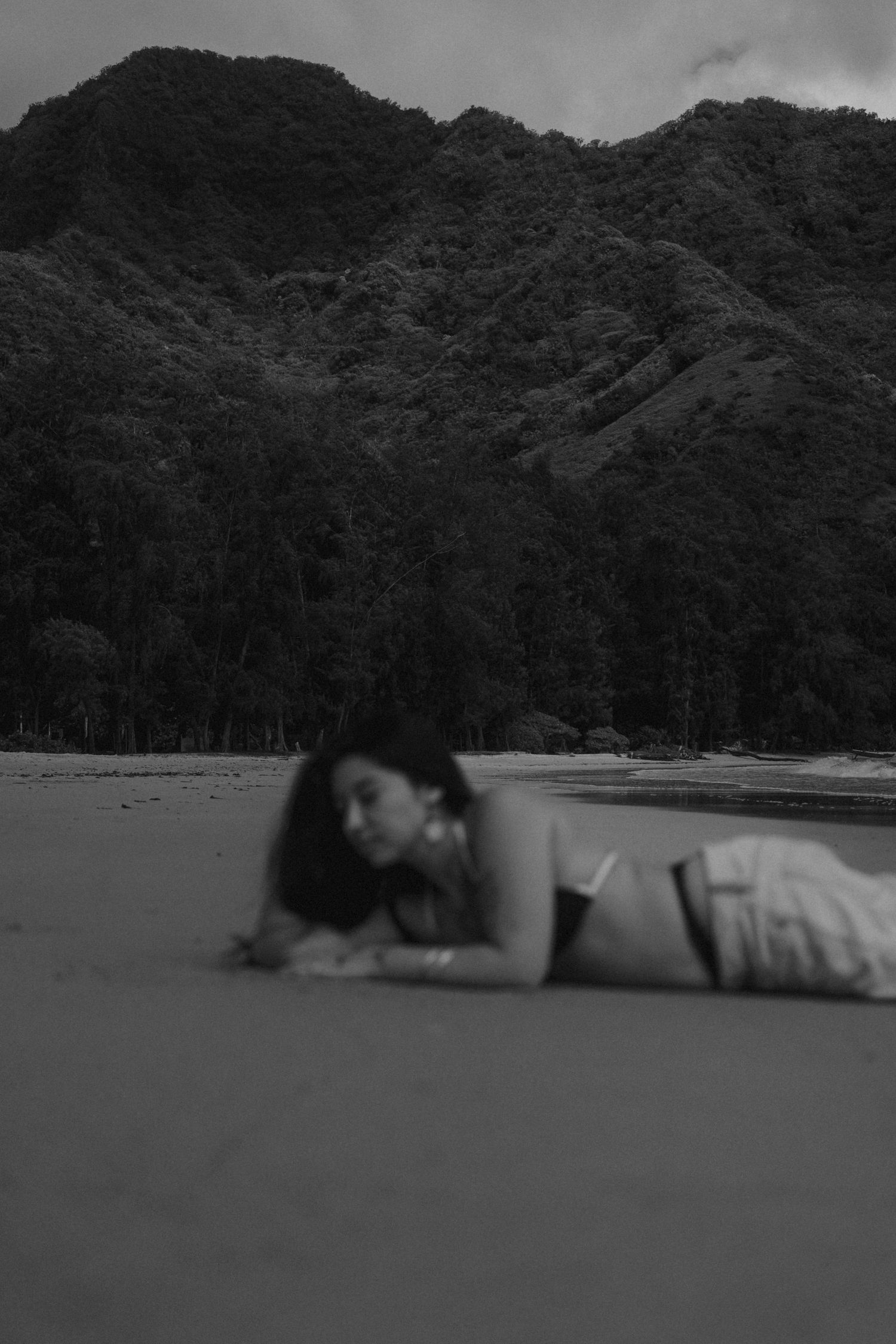 Black and white photo of woman lying on sandy beach with dramatic forested mountain backdrop.