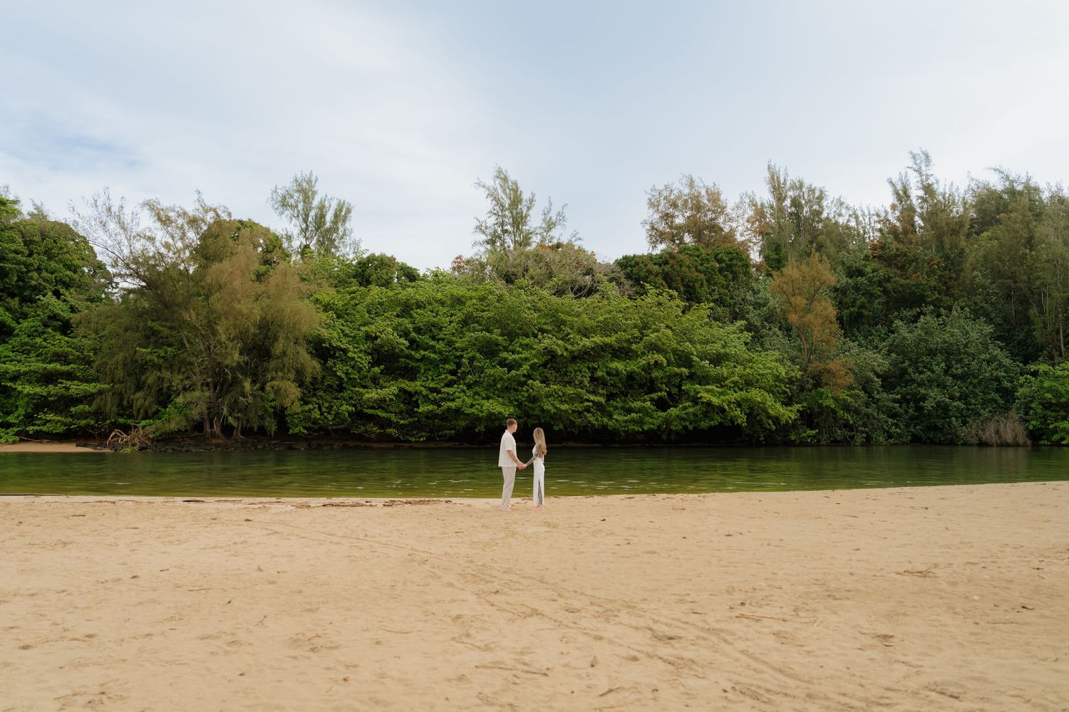 Couple holding hands on sandy beach surrounded by lush tropical greenery in Kauai Hawaii.