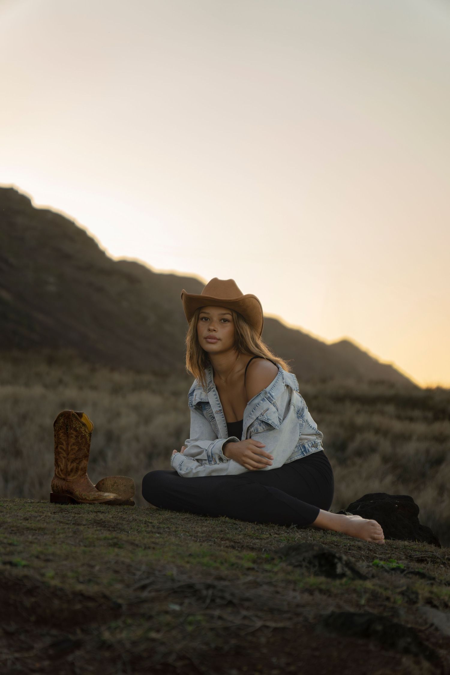 Woman in denim jacket and cowboy hat sitting cross-legged on grassy hillside at sunset with boots nearby.