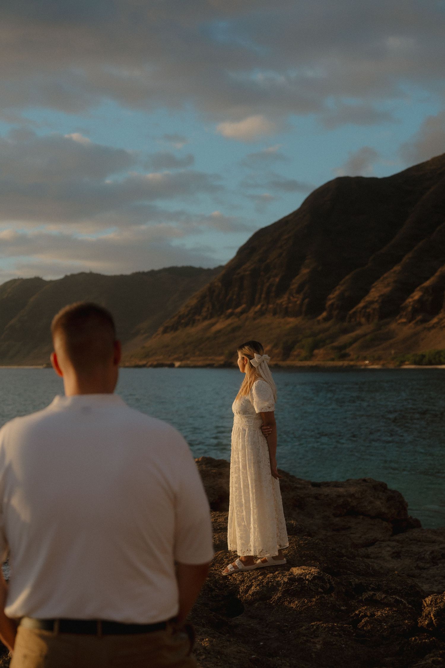 Bride in white lace dress stands on coastal rocks at sunset as groom watches nearby.