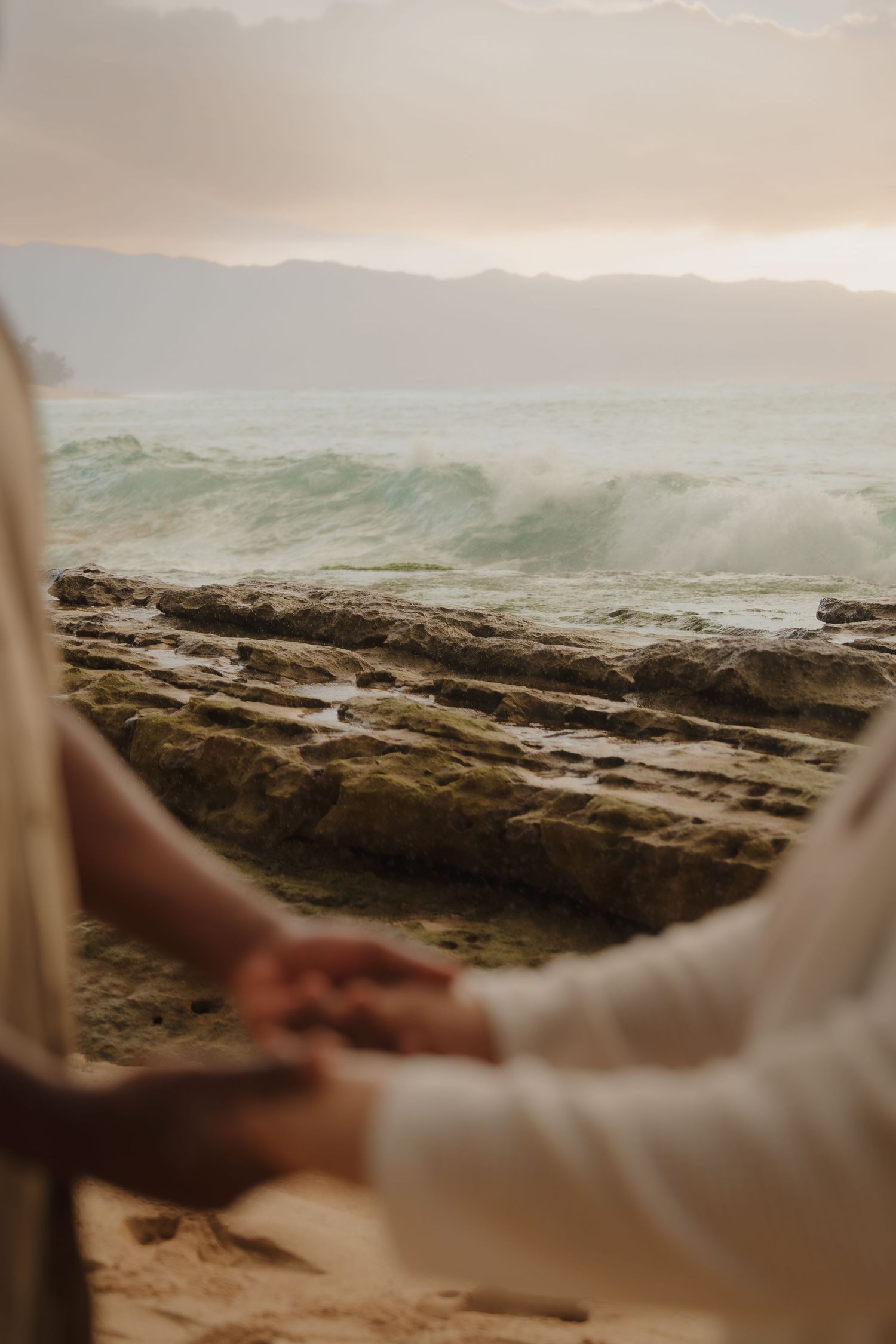 Couple holds hands on a rocky shoreline with ocean waves and mountain backdrop at sunset.