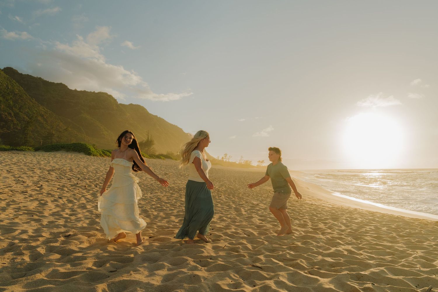 Three women hold hands dancing on a golden sandy beach at sunset with green mountains in the background.