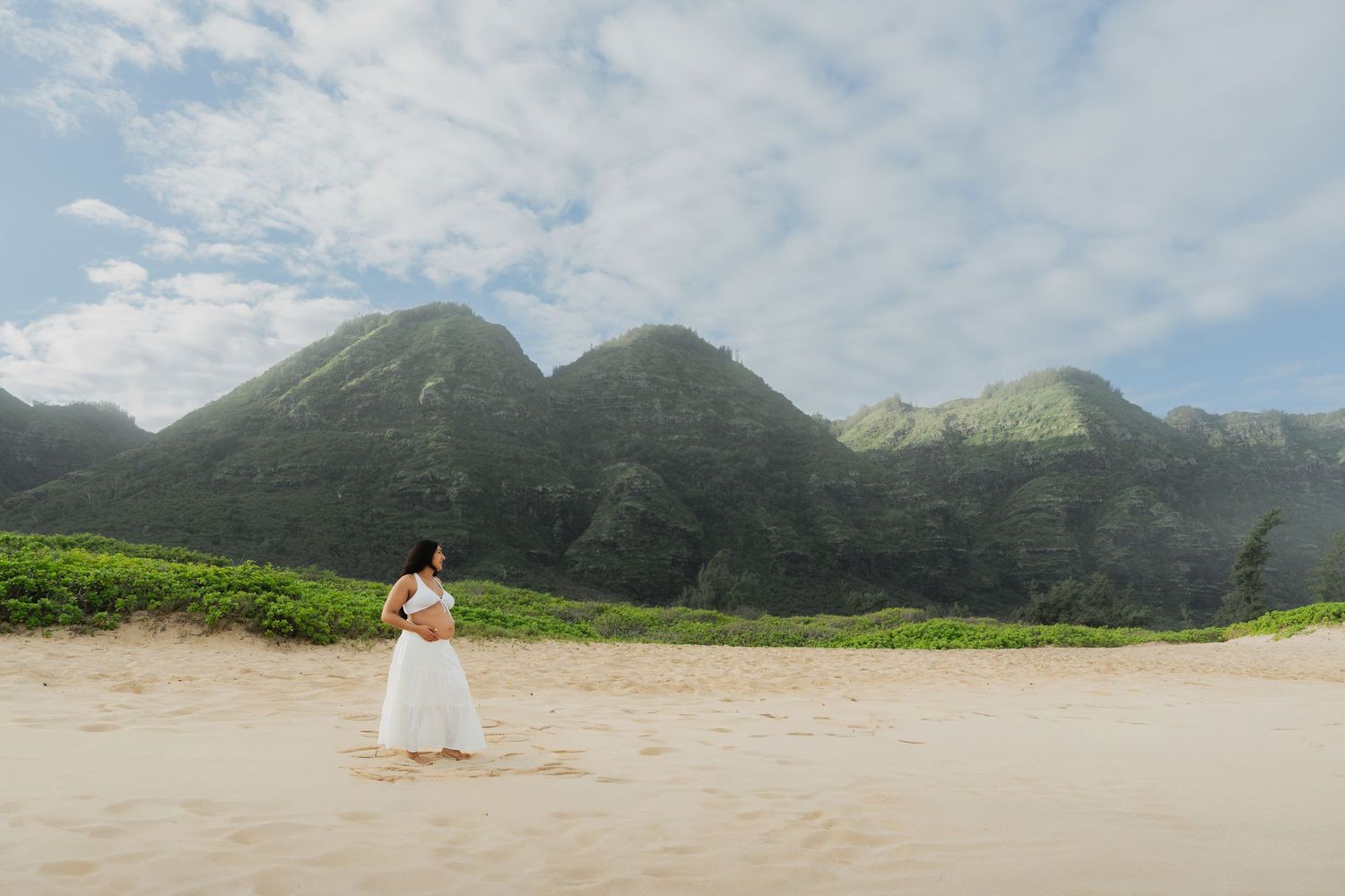 Pregnant woman in white dress standing on beach with lush green mountains behind.