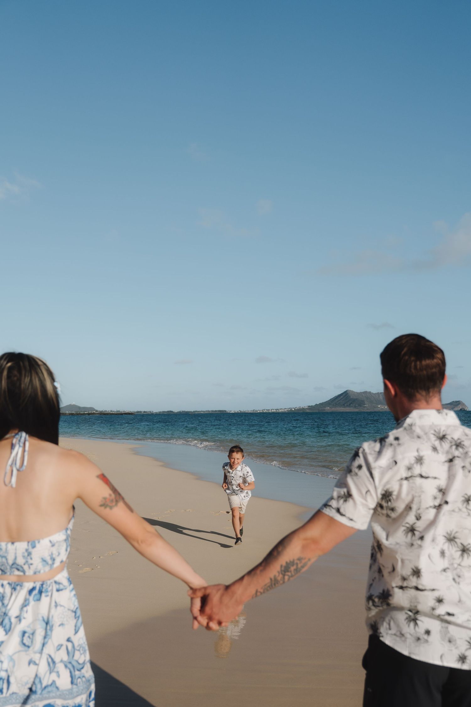 Couple holding hands on a sandy beach while a young boy runs toward them with ocean backdrop.