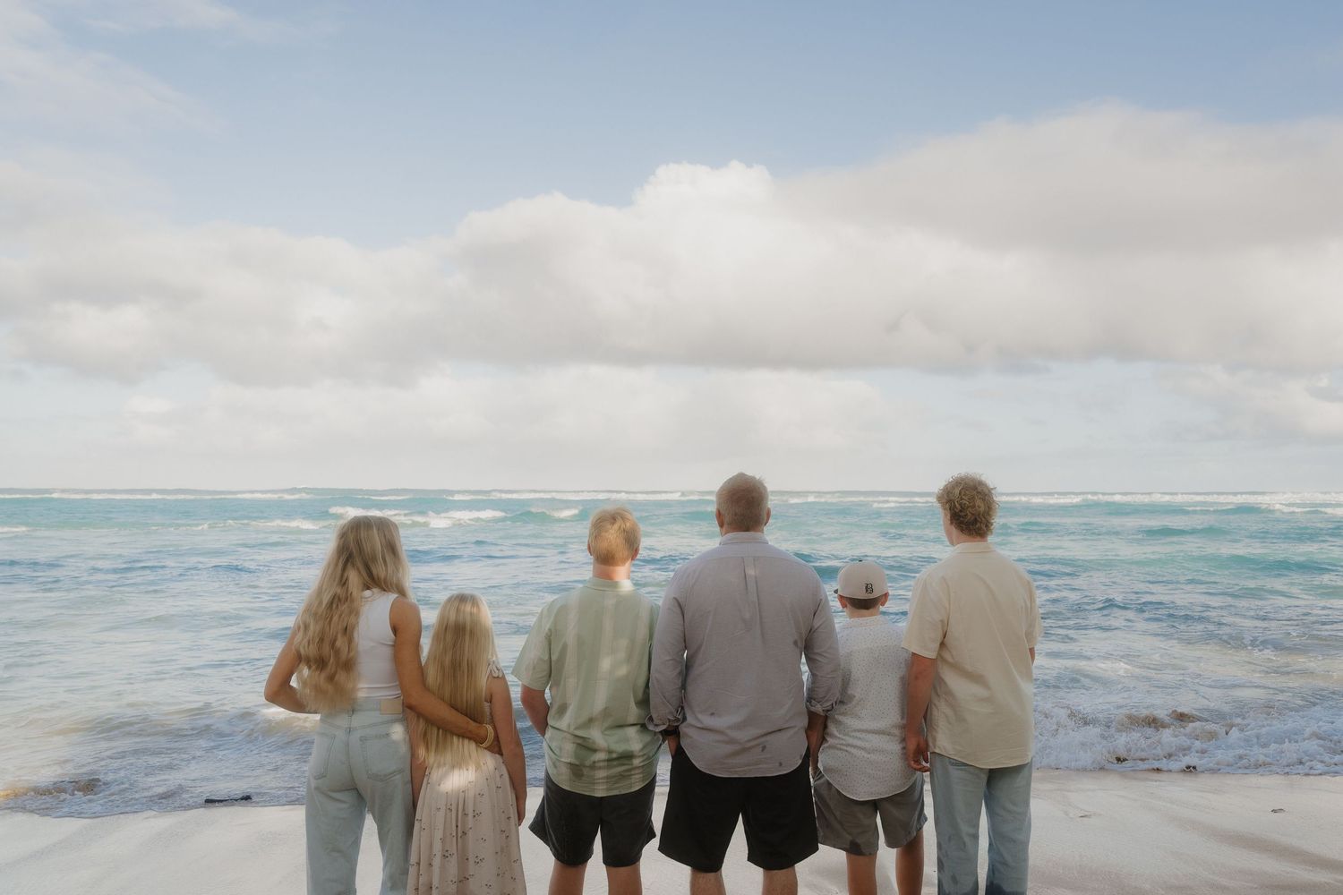 Family of six standing with backs to camera gazing at ocean waves on sunny beach.