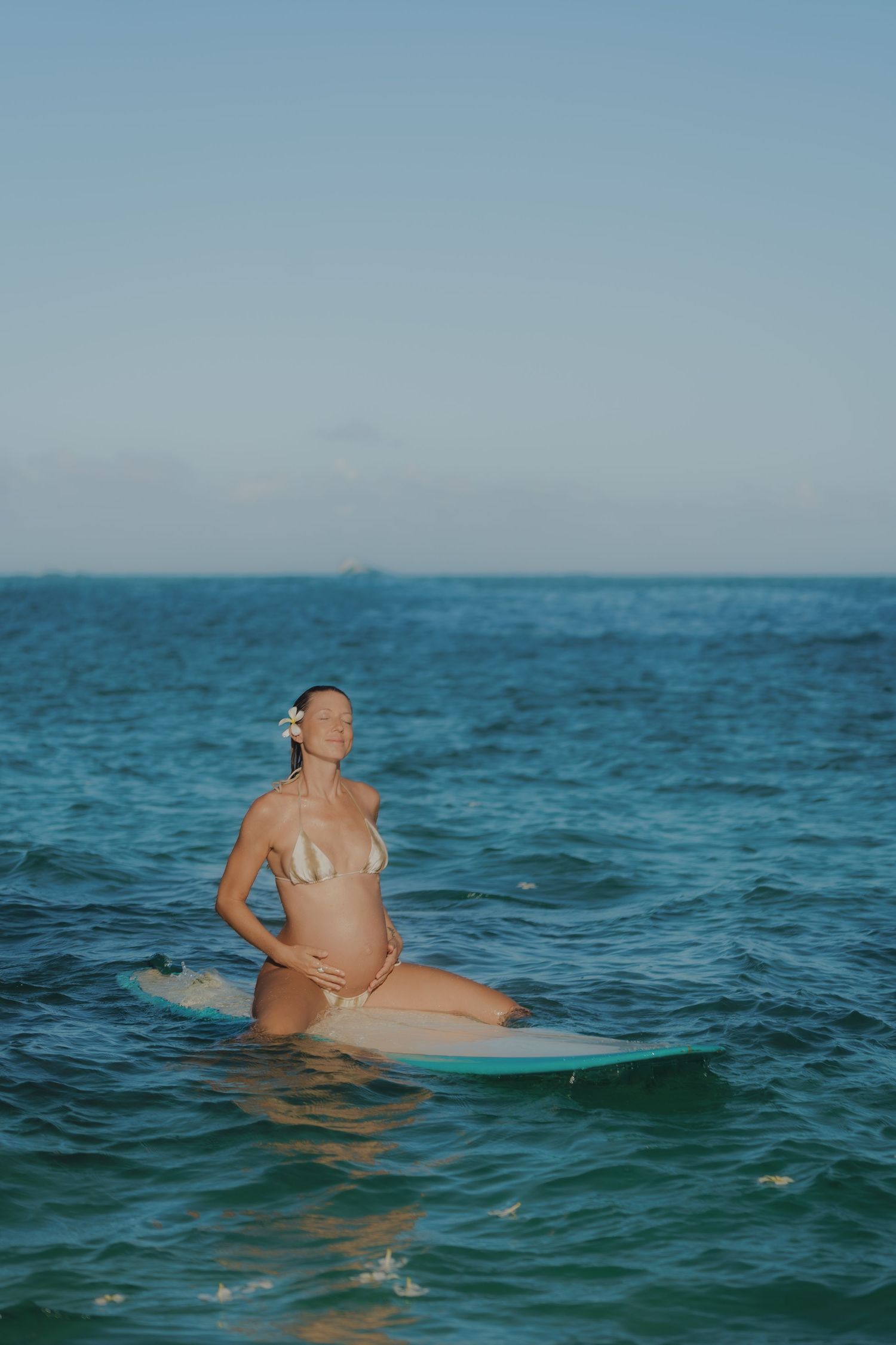 Pregnant woman in bikini sitting on surfboard in calm turquoise ocean water.