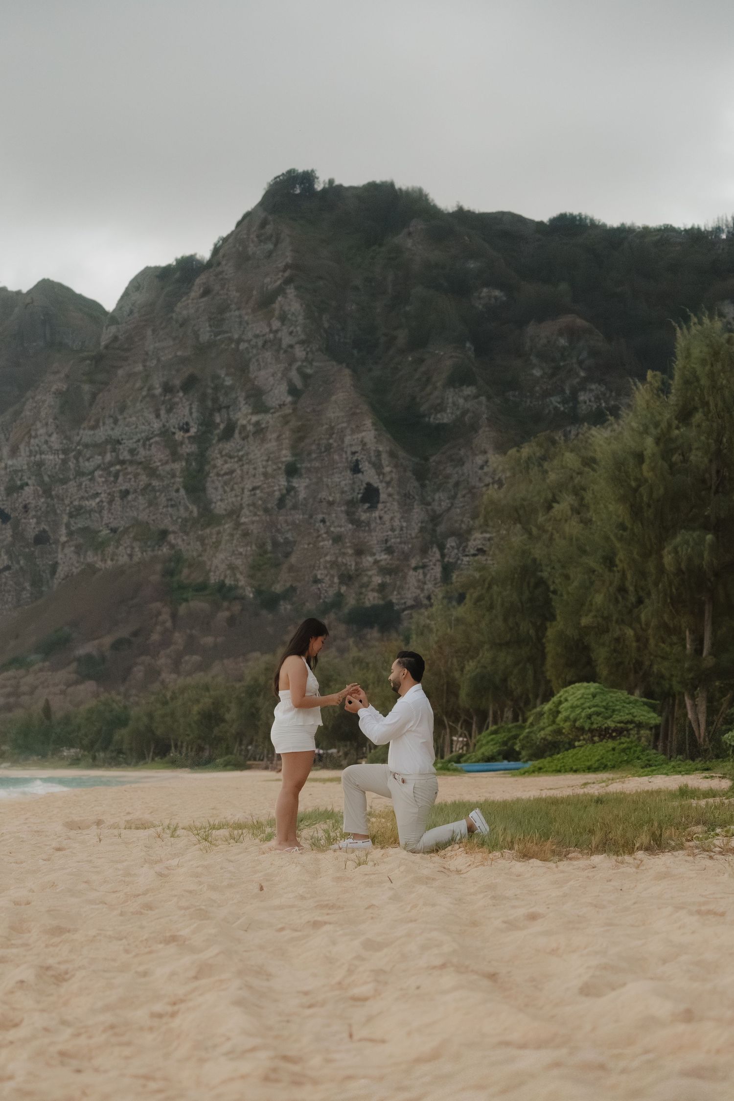 Man proposes on one knee to woman on a sandy beach with lush mountain cliffs behind them.