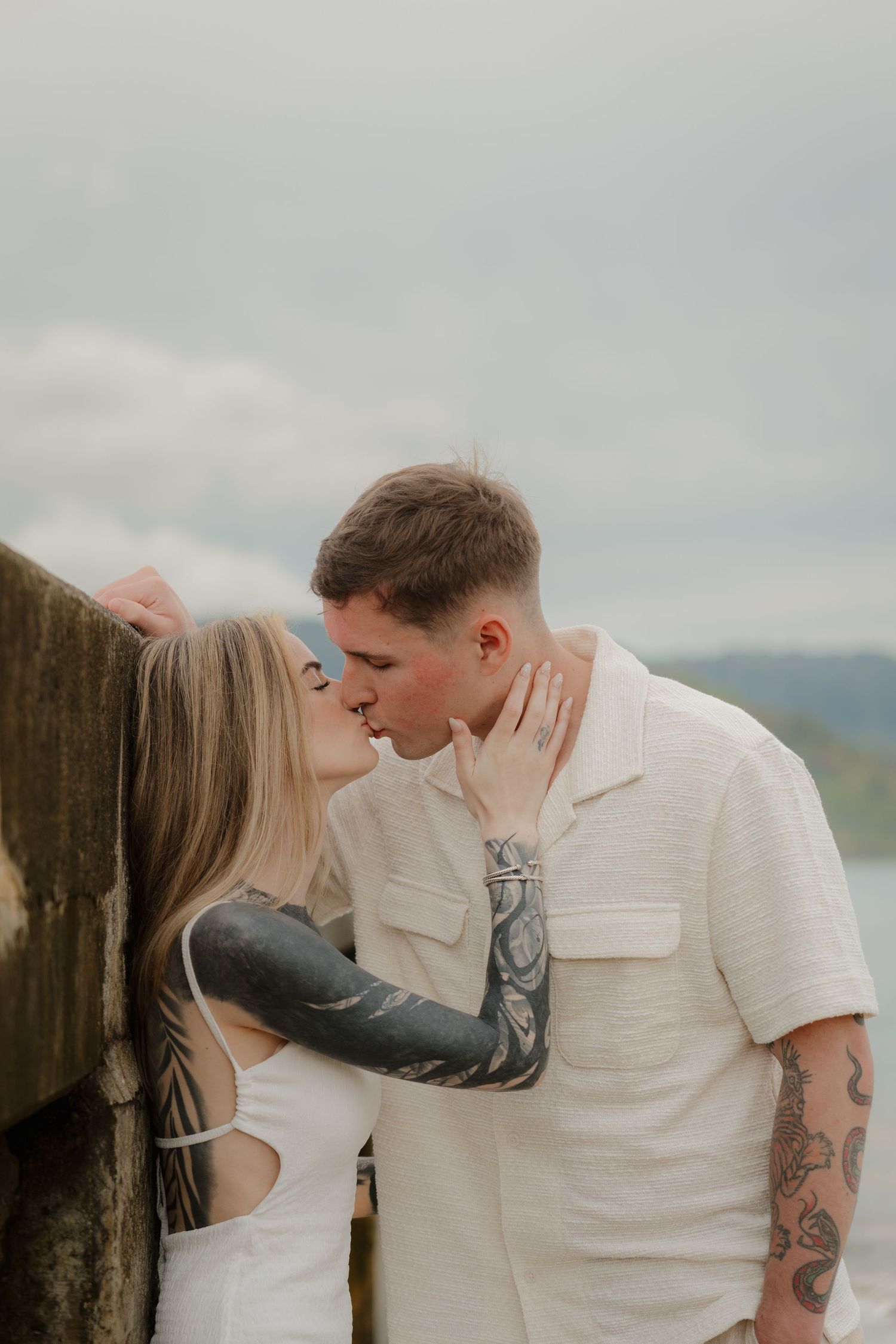 Tattooed couple sharing a romantic kiss outdoors with cloudy sky backdrop.
