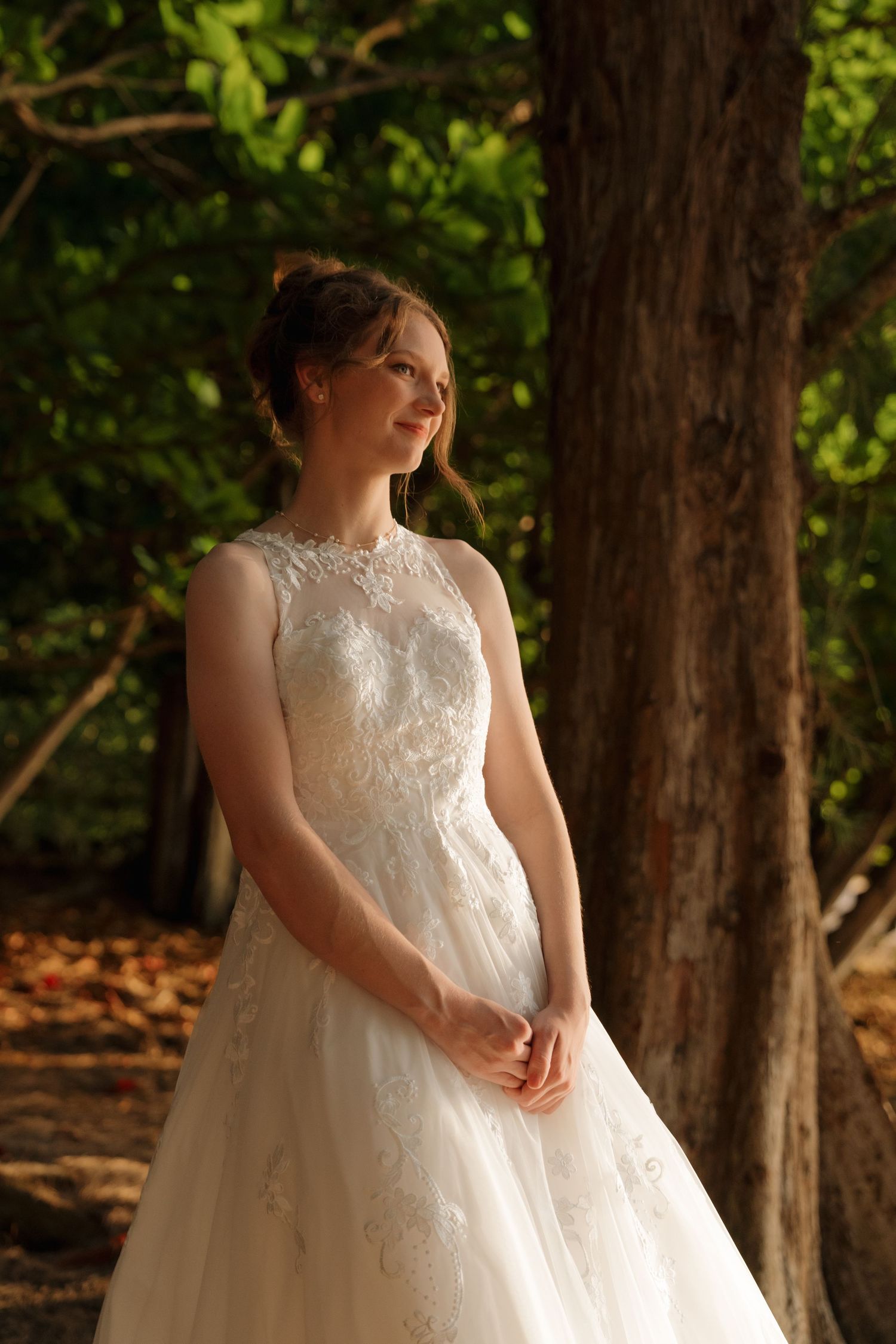 Elegant bride in a lace wedding gown standing near a tree in a sunlit forest setting.